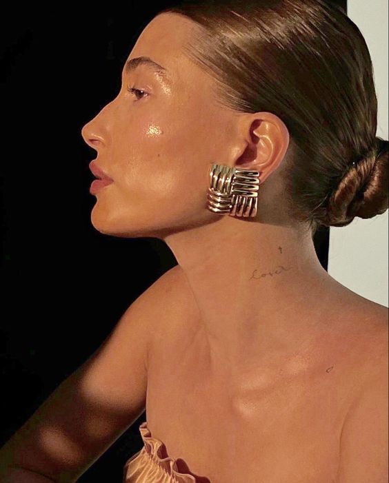 A woman with slicked-back hair in a bun is shown in profile. She wears large, gold, geometric earrings and a strapless top. Soft light highlights her glowing skin and a small love tattoo on her neck.