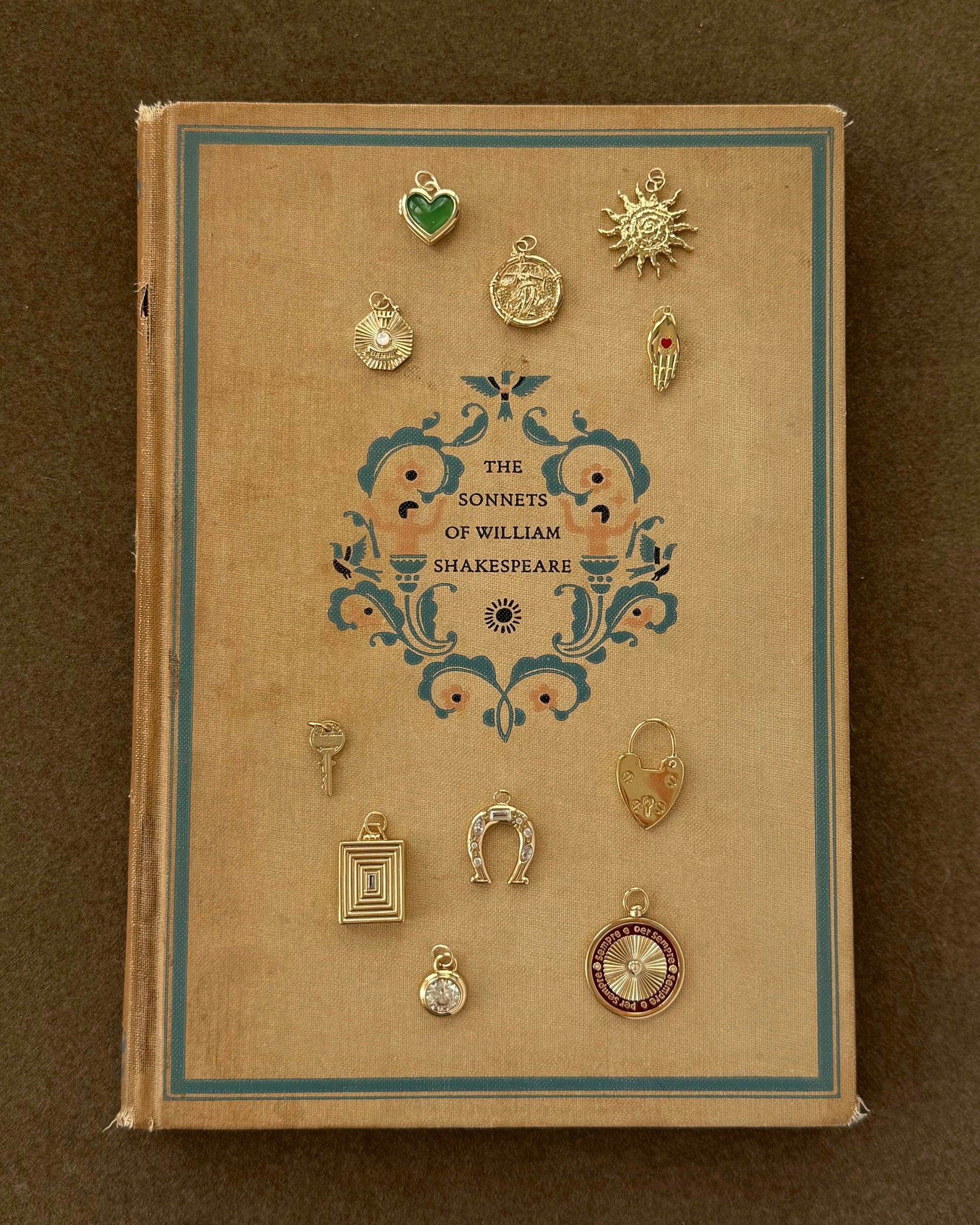 A vintage book titled "The Sonnets of William Shakespeare" rests on a brown surface. Twelve charms, including the Mod + Jo Davie Heart Birthstone Locket Charm with genuine gemstones, are arranged around the ornate cover.