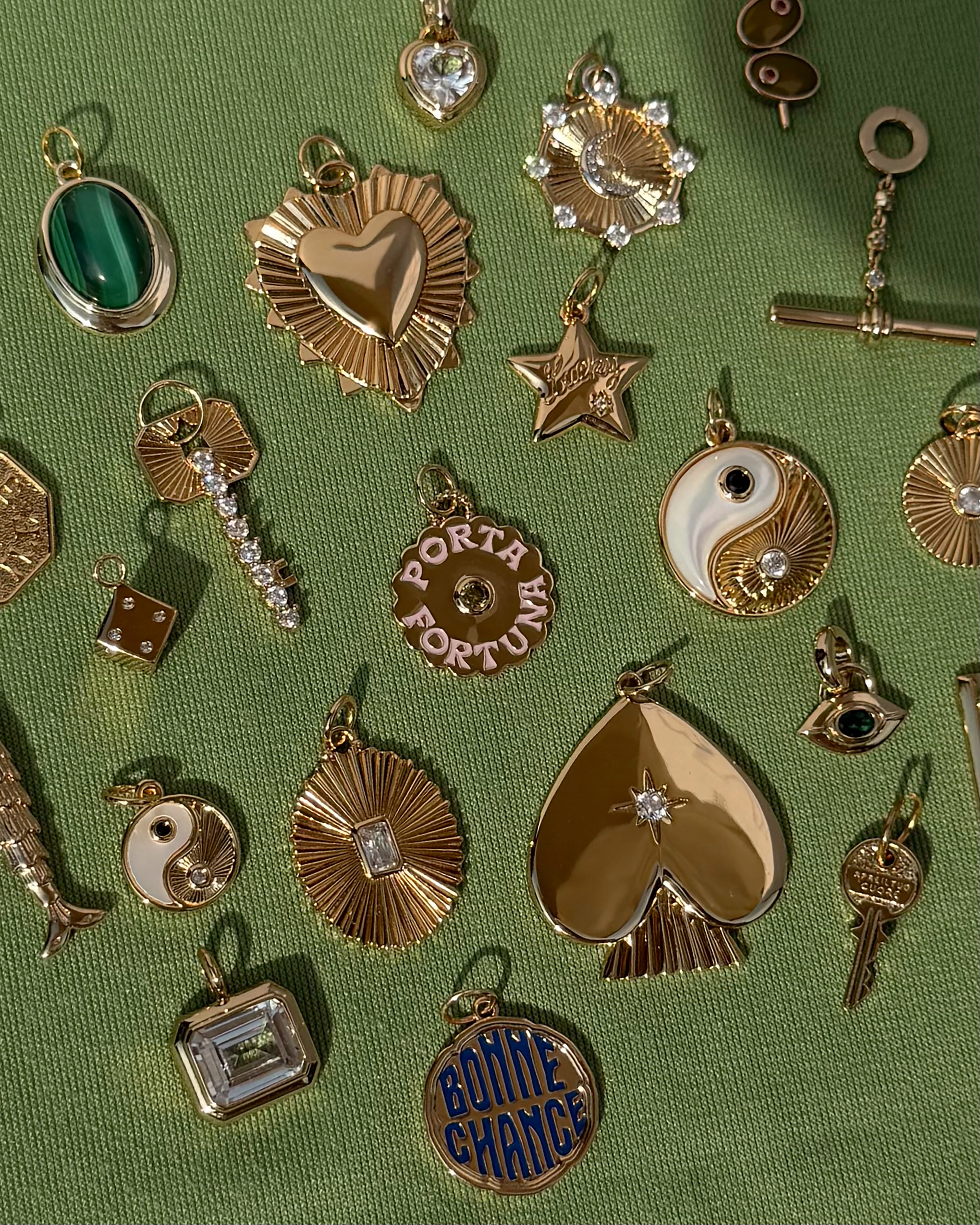 Assorted gold and gemstone charms—including hearts, stars, dice, yin yangs, and a “BONNE CHANCE” pendant—are artfully displayed on textured green fabric alongside a sparkling emerald-cut crystal. Among them is the Cora Fluted Charm by Mod + Jo, an elegant piece crafted in radiant 14k gold for timeless appeal.