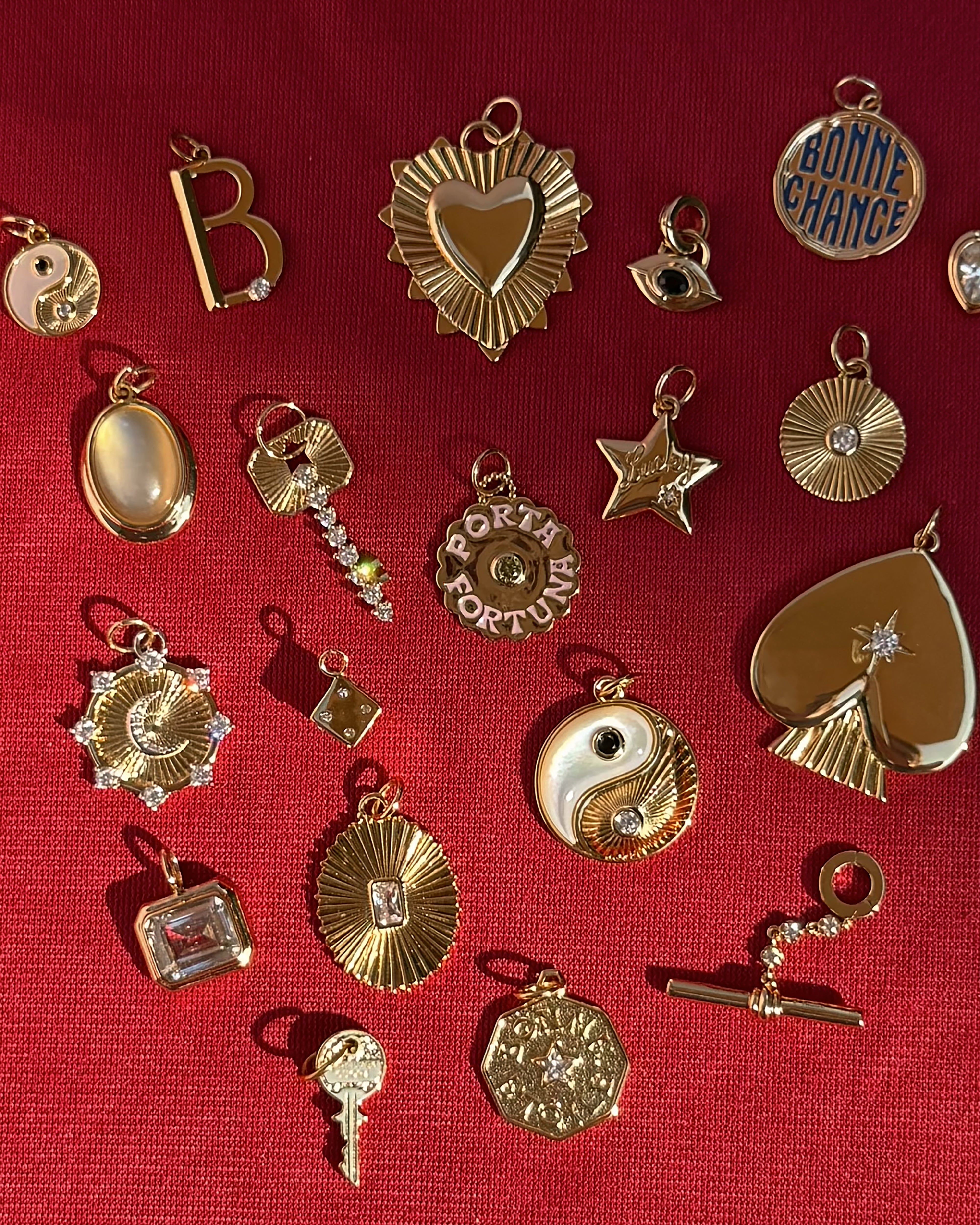 A variety of gold and gemstone charms—including hearts, stars, a key, yin-yang, lucky charm motifs, as well as the Mod + Jo Porta Fortuna Charm and Bonnie Chance medallions—are artfully arranged on a textured red surface next to an elegant 14k gold charm and a striking gold coin necklace.