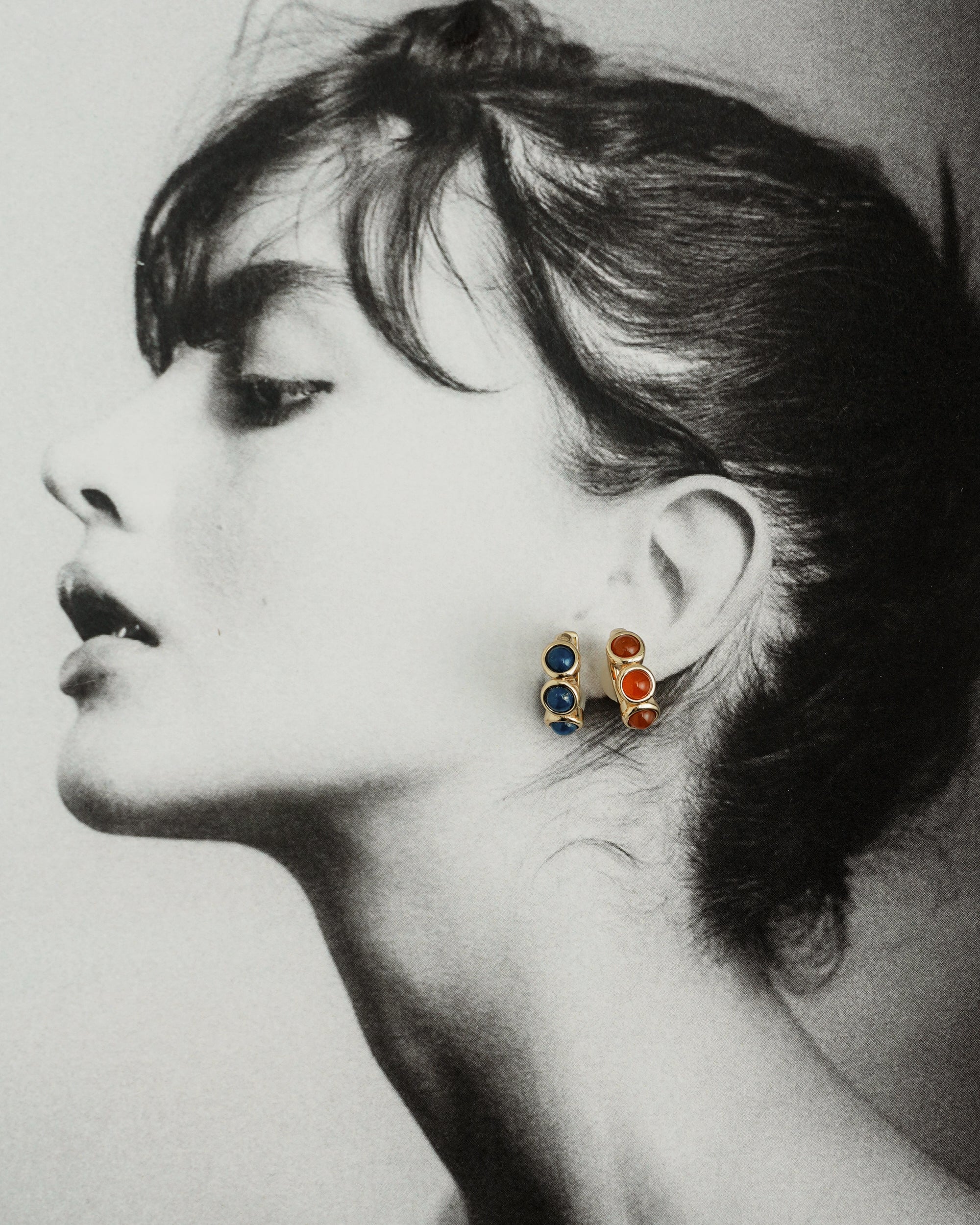 Black-and-white profile photo of a woman with an elegant updo, showcasing Mod + Jo’s *Preorder* Corynn Gemstone Huggies in blue and red stones. The image highlights her graceful neck and facial features.