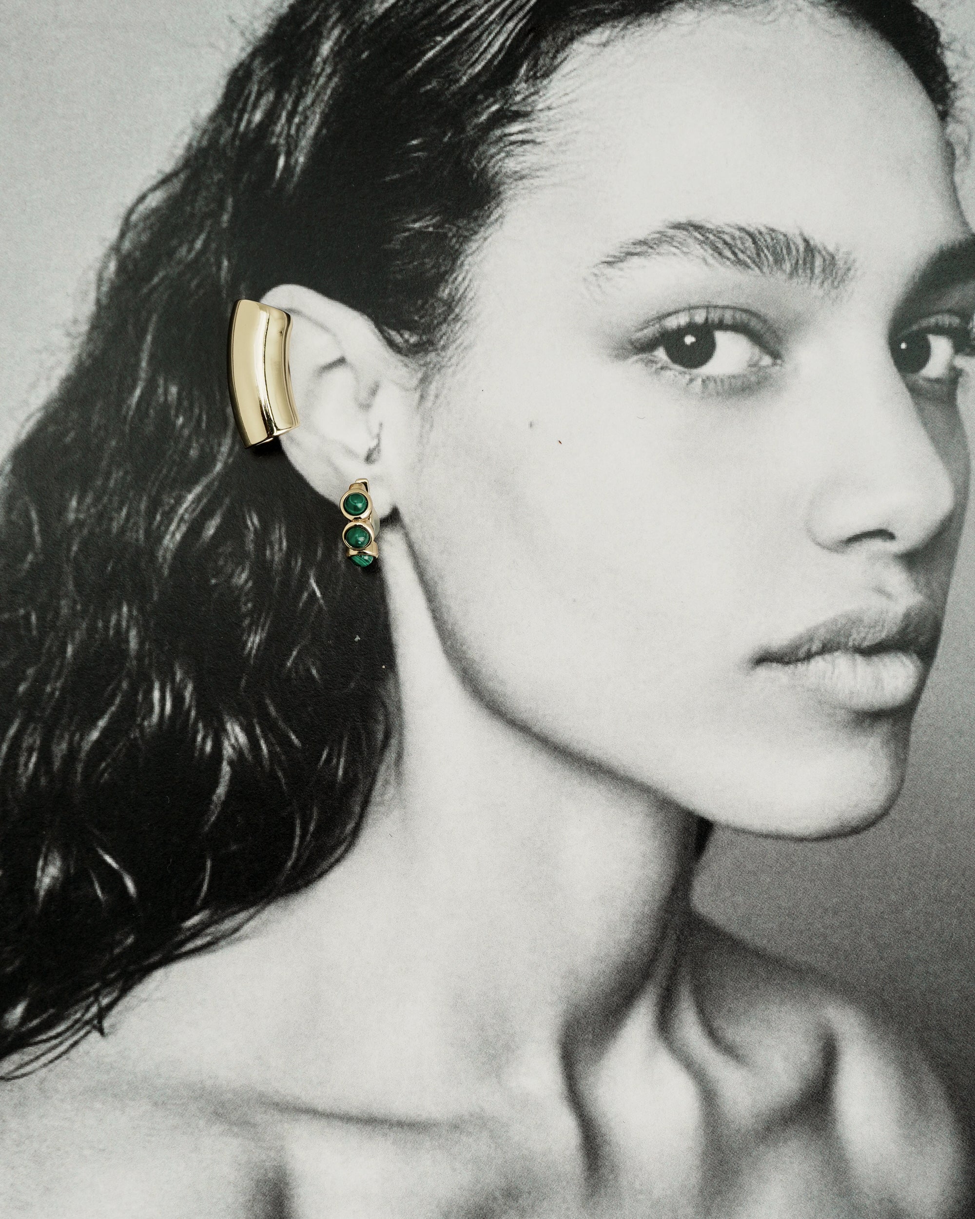 Black and white portrait of a woman with wavy hair wearing Mod + Jo’s *Preorder* Corynn Gemstone Huggies—gold ear cuffs with two dangling green gemstones. She gazes calmly to the side.