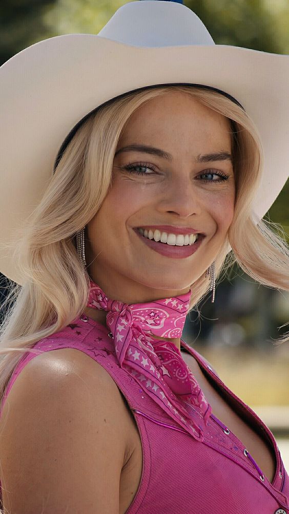 A woman with long blonde hair, wearing a white cowboy hat, pink neck scarf with star patterns, and a pink top, smiles brightly outdoors.