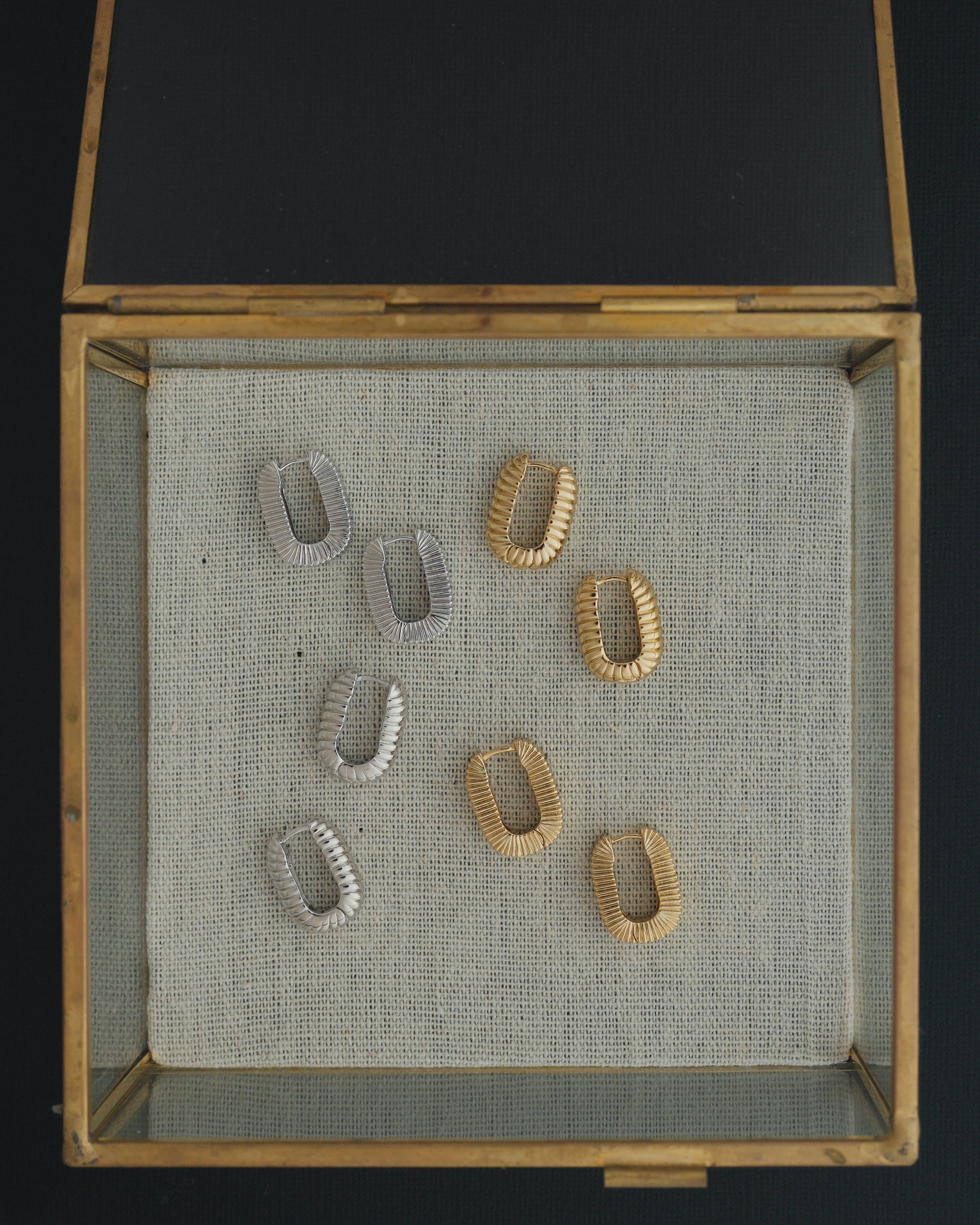 A glass and brass display box features four pairs of Mod + Jo Elly French Rope Hoops—including two shimmering gold and two silver pairs—beautifully arranged on a textured beige fabric background. These elegant earrings combine timeless style with lightweight versatility, making them perfect for those who love the classic look of hoop earrings.