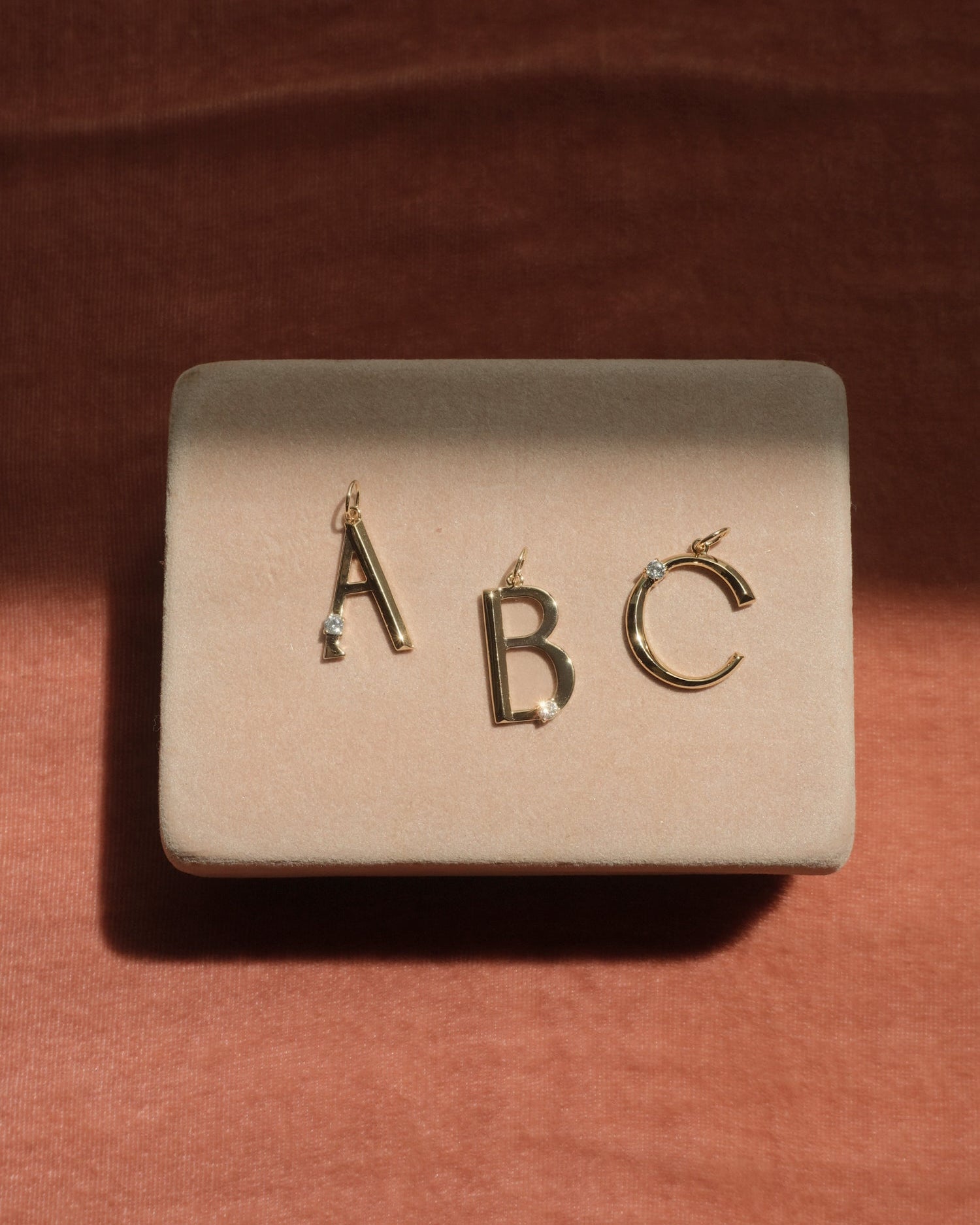 Mod + Jo’s Eloise Letter Charm pendants shaped as A, B, and C are showcased on a beige velvet stand with a rust-colored fabric background.