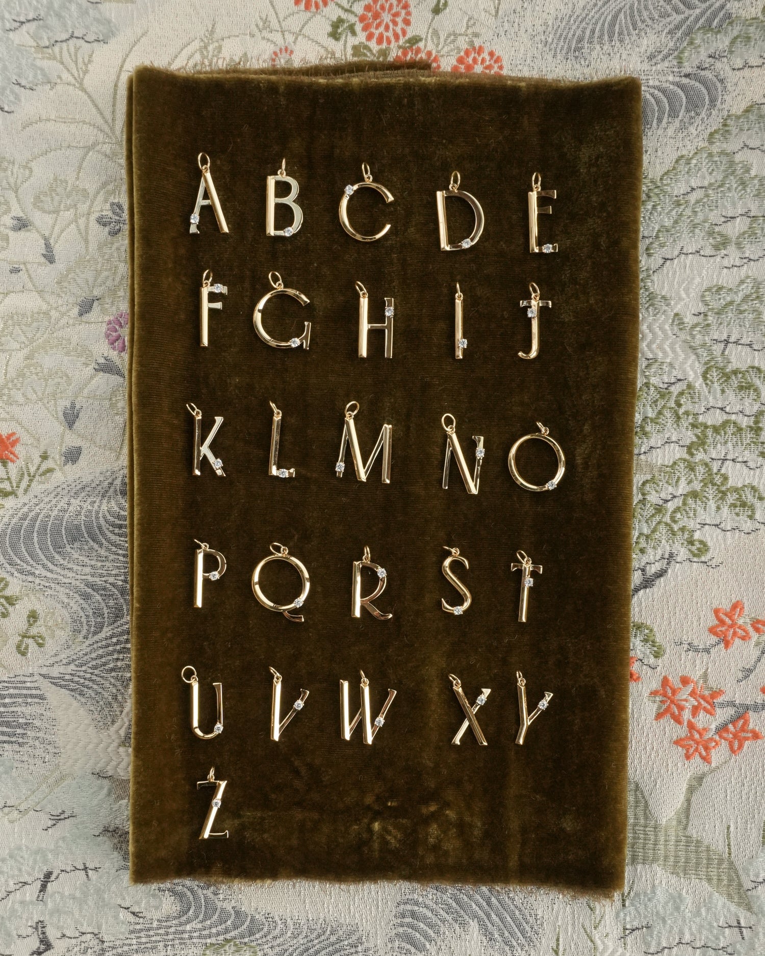 A set of Eloise Letter Charms by Mod + Jo, gold plated and arranged in four rows on a dark velvet board, displayed over patterned fabric with floral and wave designs.