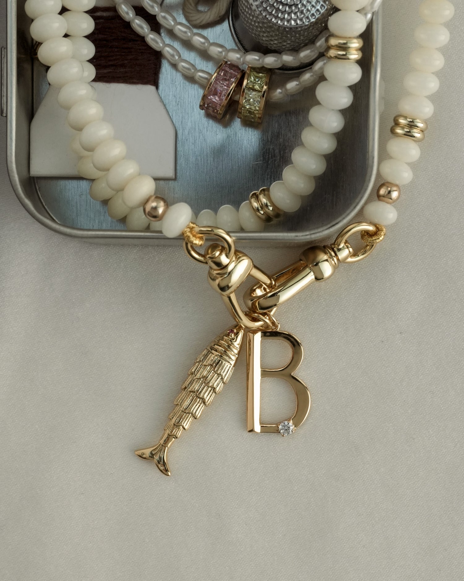 A close-up of beaded jewelry and rings on a metal tray, featuring a gold plated necklace with a fish charm and Mod + Jo’s Eloise Letter Charm—a letter B pendant adorned with a small gem.