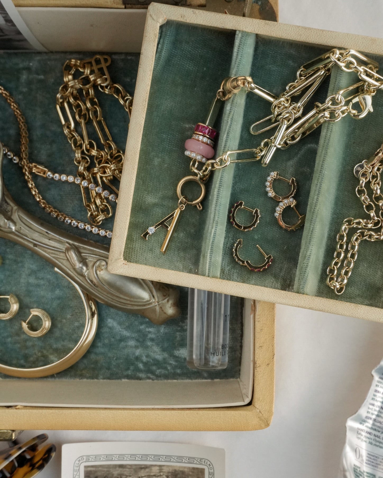 An open jewelry box with Mod + Jo's Harlow Reversible Birthstone Huggies, gold chains, a beaded pendant, and a bangle on soft green lining. Gold-plated earrings and a card sit next to a clear tube.