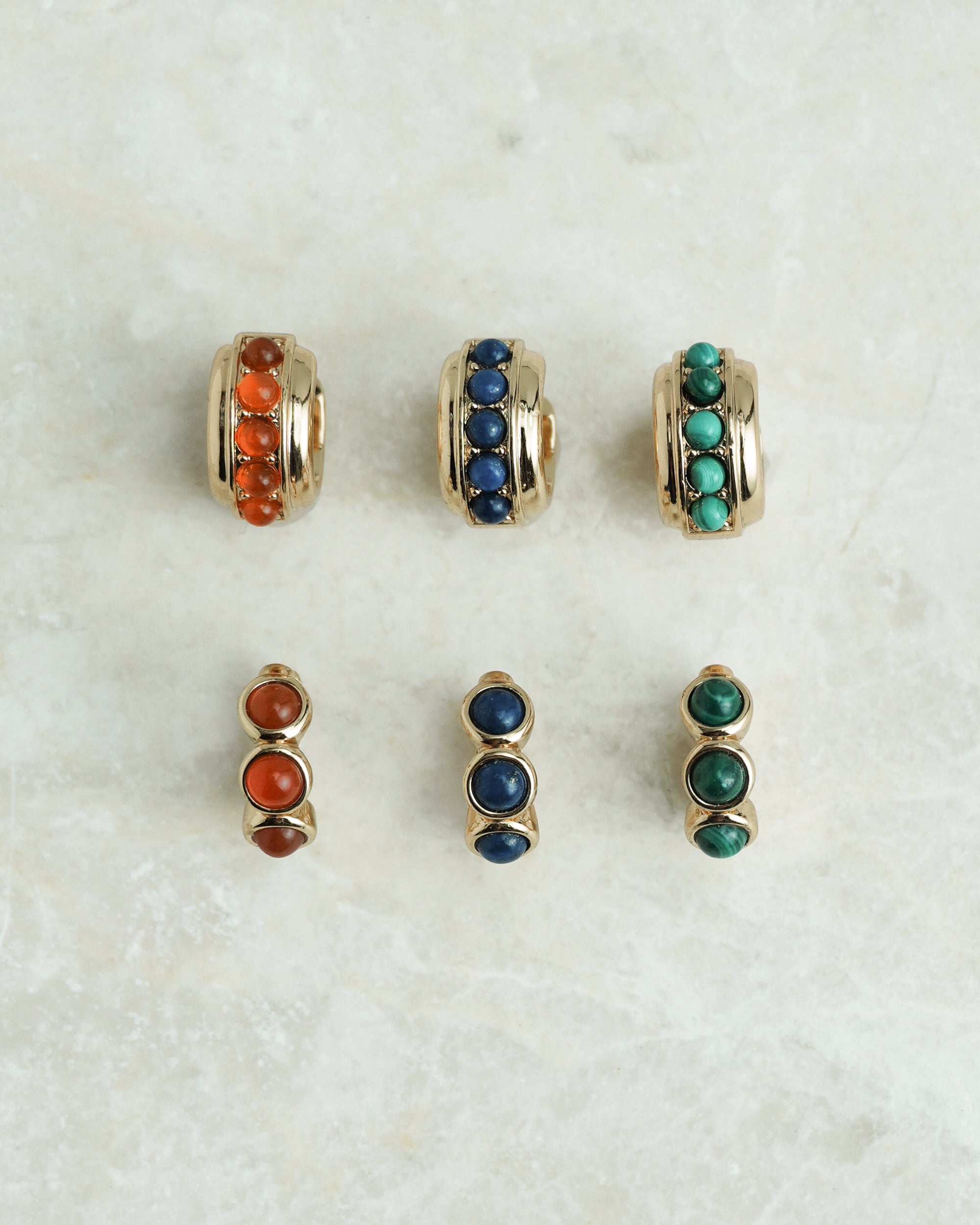 Six pairs of *Preorder* Corynn Gemstone Huggies by Mod + Jo—18k gold plated earrings with round red, blue, and green stones—are shown on a marbled surface; each color has three-stone hoops and two-stone studs.