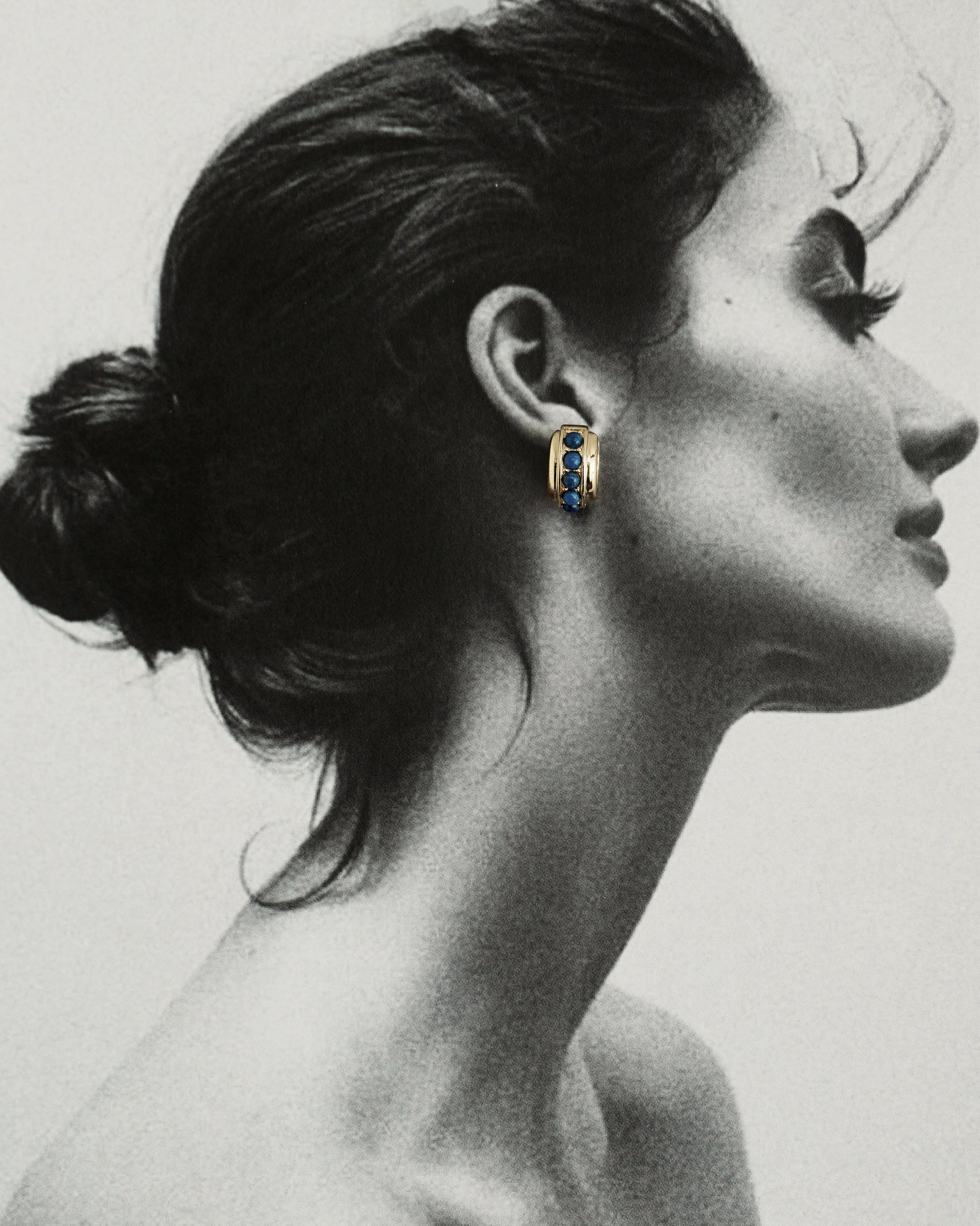 A black-and-white profile photo of a woman with her hair in a bun, wearing Mod + Jo’s *Preorder* Evy Raised Gemstone Hoops. The genuine lapis lazuli gem is shown in color, standing out against the monochrome image.