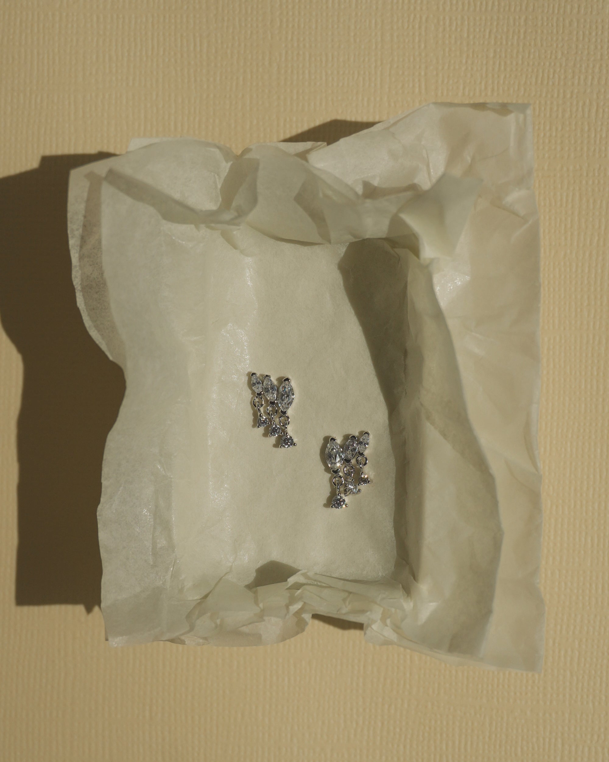 A pair of Mod + Jo Gabby Marquise Dangle Studs, featuring small, silver, flower-shaped cubic zirconia settings, rests on crumpled tissue paper against a textured, pale yellow background.