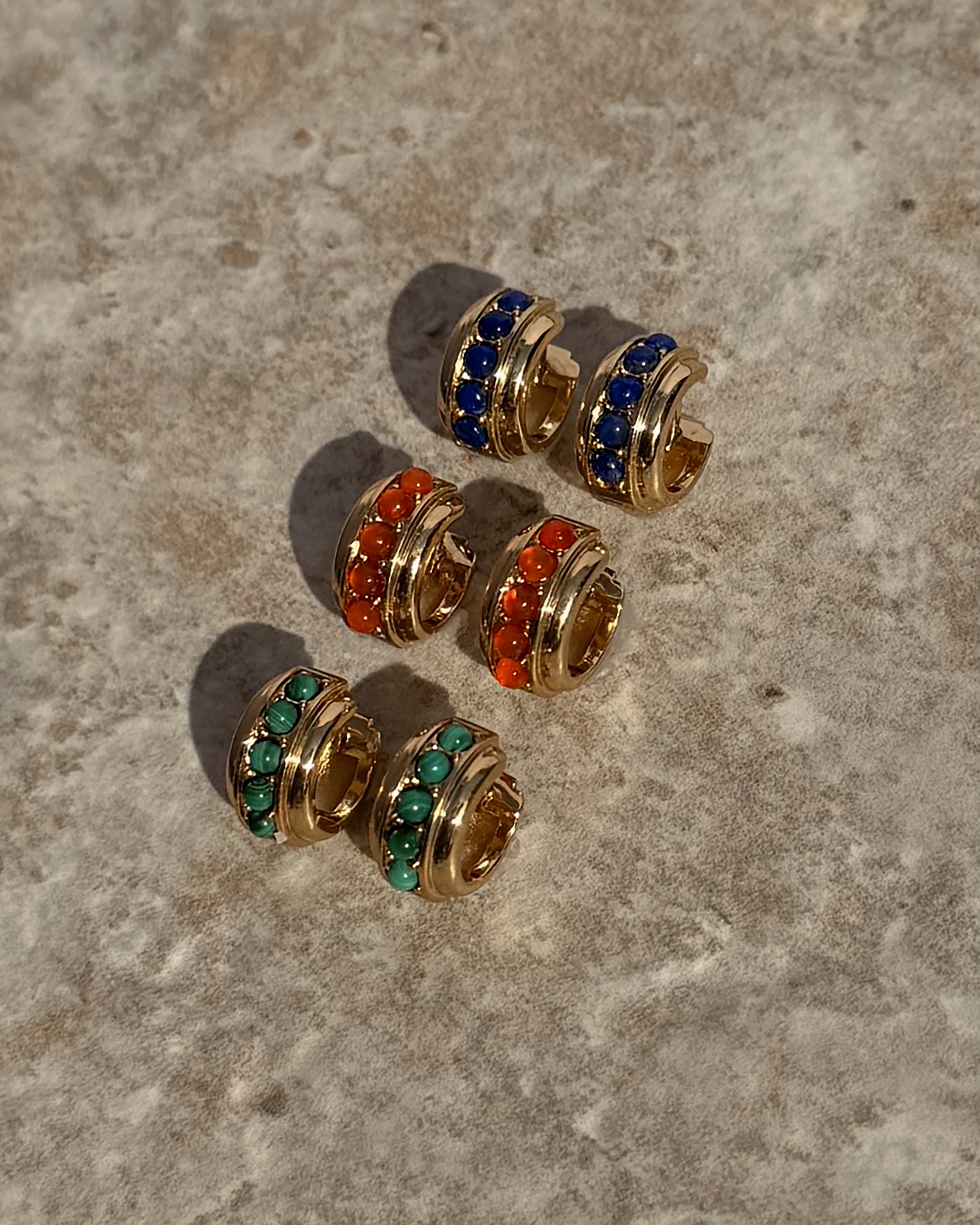Three pairs of Mod + Jo *Preorder* Evy Raised Gemstone Hoops, each 18k gold plated and set with tiny blue, red-orange, and green stones, are displayed on a textured stone surface.