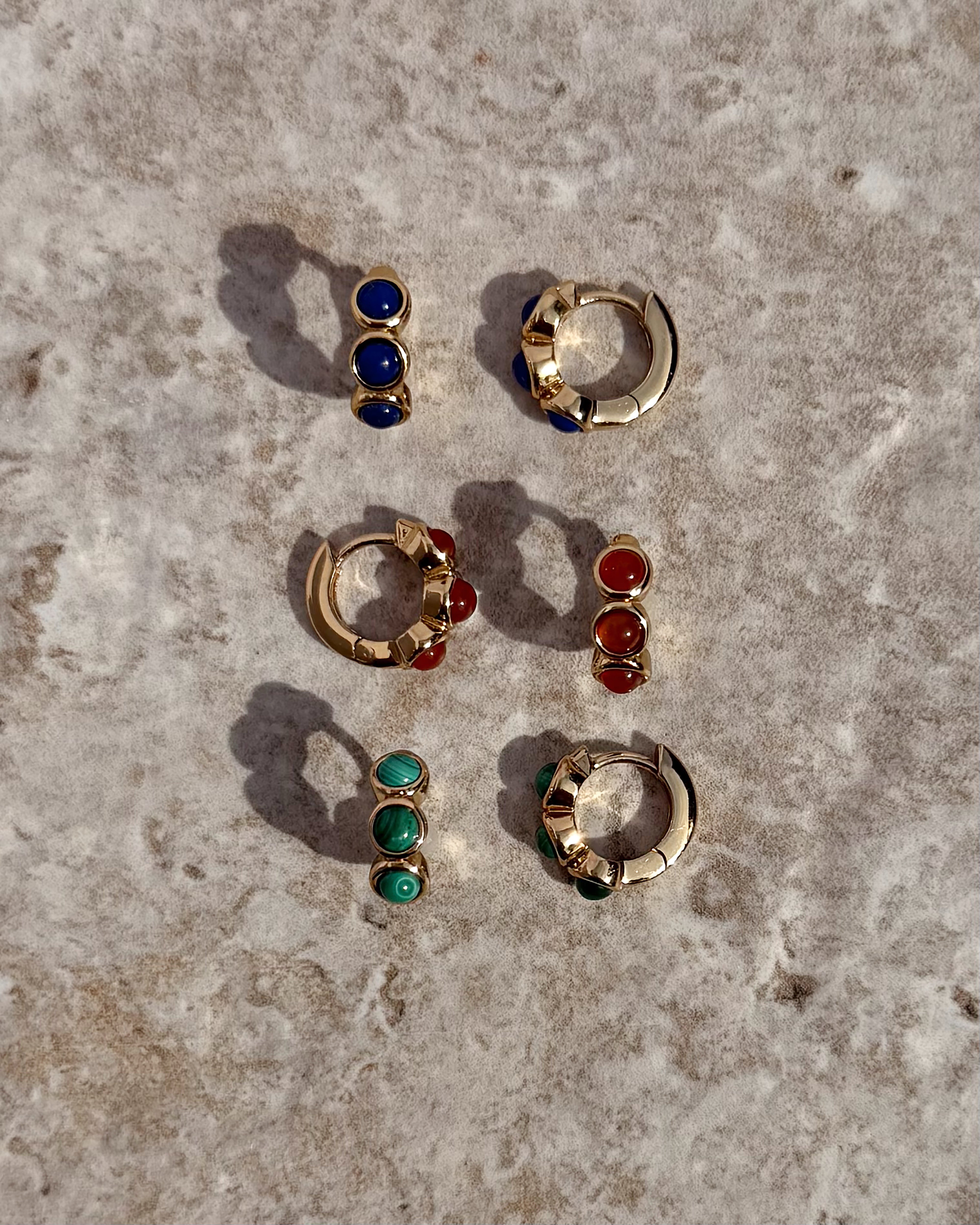 Three pairs of Mod + Jo *Preorder* Corynn Gemstone Huggies—gold earrings with blue, red, and green stones—are arranged on textured stone. Each 18k plated set includes a hoop and matching two-stone stud for versatile wear.