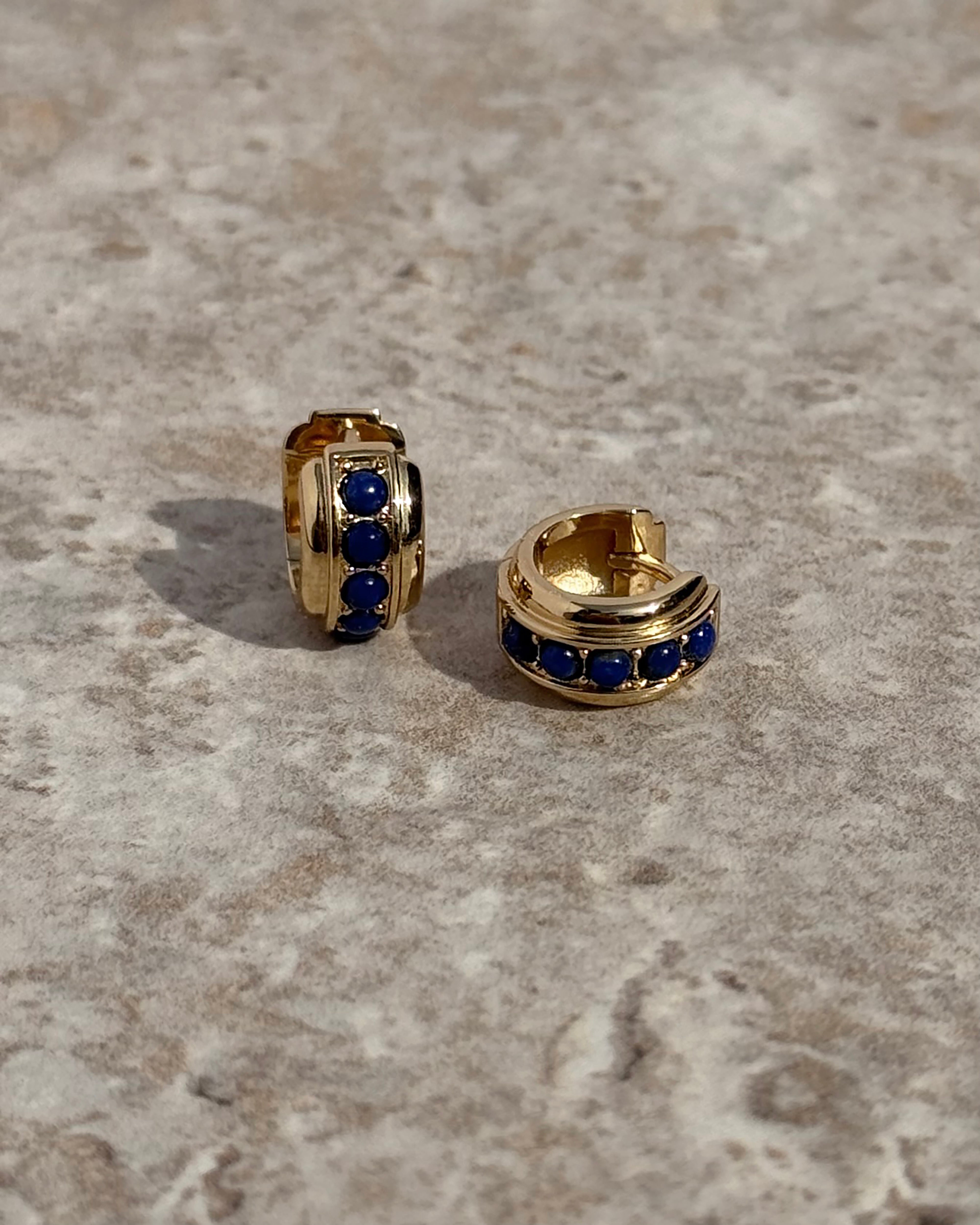 The *Preorder* Evy Raised Gemstone Hoops by Mod + Jo feature small gold hoops, each adorned with three genuine lapis lazuli stones, showcased on a lightly textured surface.