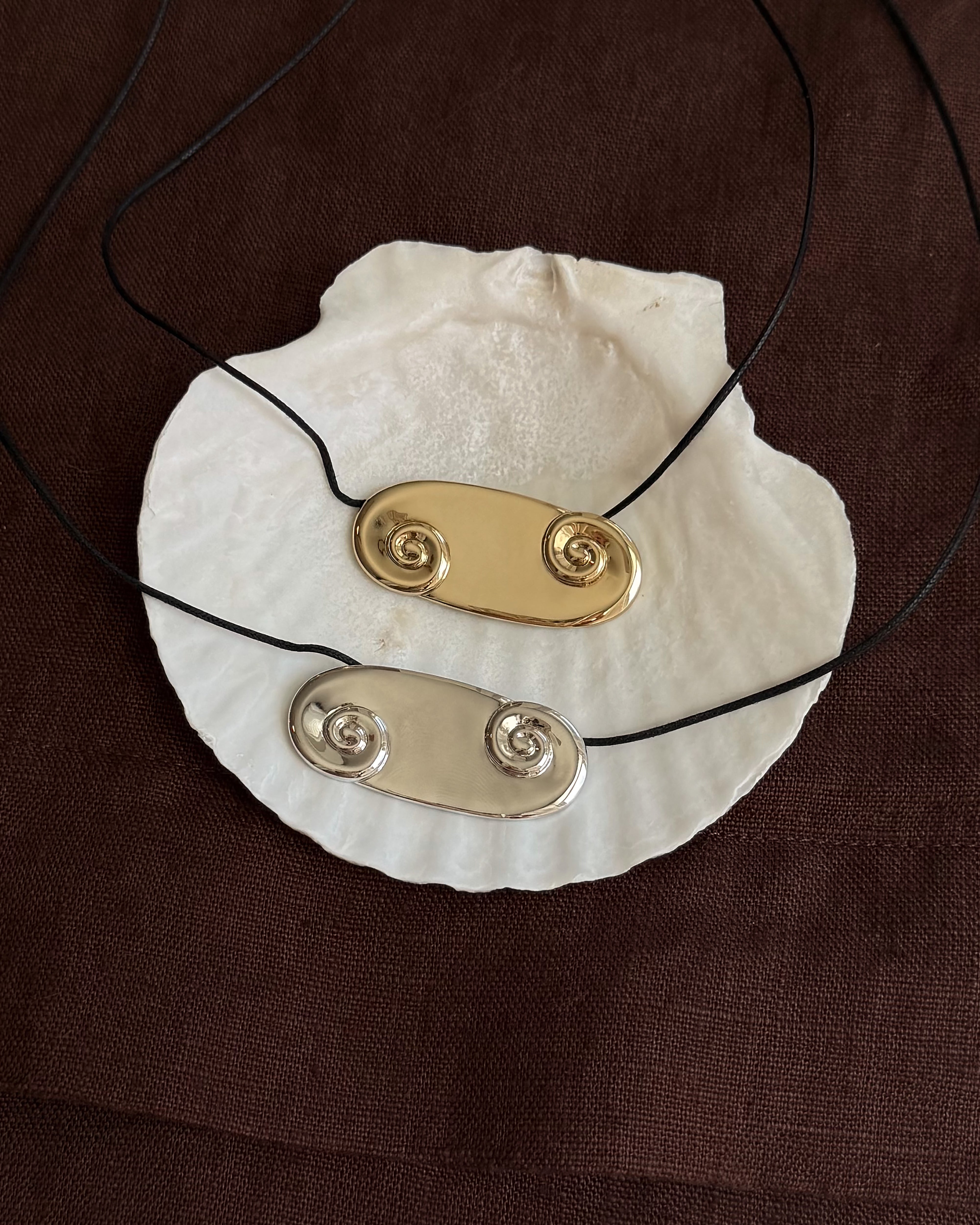 The Suna Spiral Cord Necklaces by Mod + Jo, in gold and silver oval double-spiral designs, are shown on a white seashell atop brown fabric. Each pendant dangles elegantly from a black cord necklace.