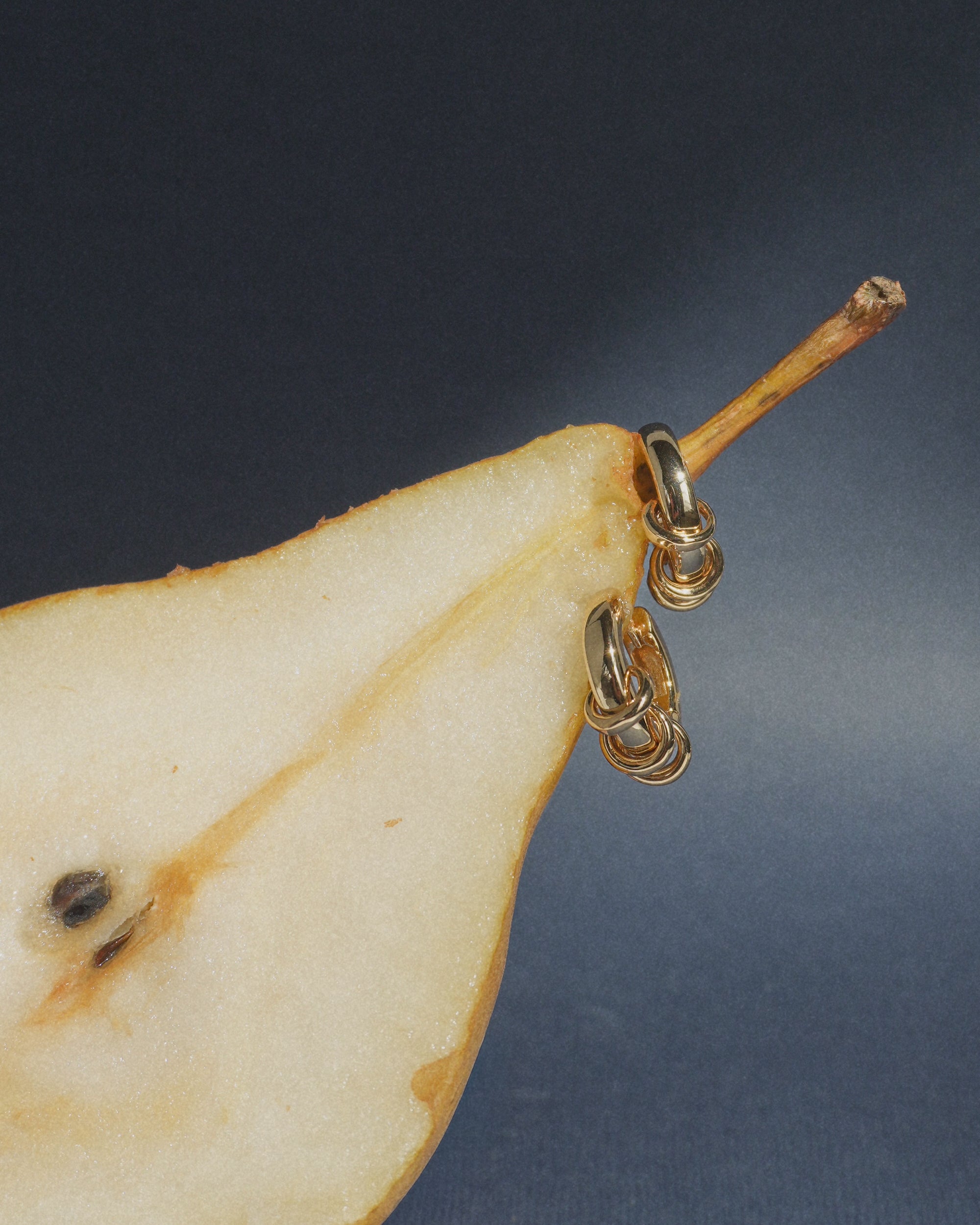 The Grande Levi Huggies by Mod + Jo are displayed hanging on the stem of a sliced pear, set against a dark blue background—a subtle showcase of timeless style and hypoallergenic jewelry.