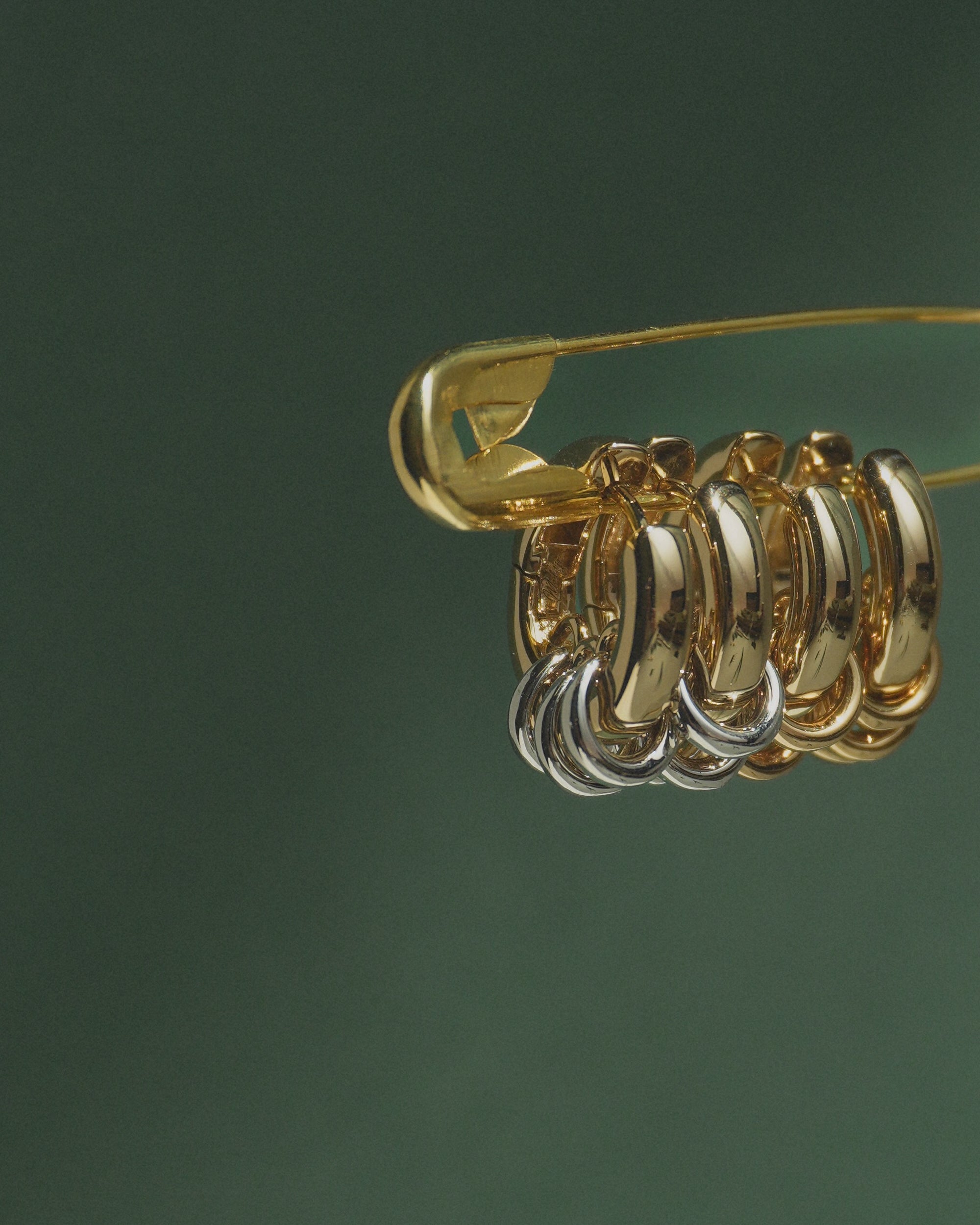 A gold safety pin displays several metallic hoop earrings—primarily gold with a touch of silver—against a dark green background, featuring the Mod + Jo Grande Levi Huggies for an elevated, statement-making style.
