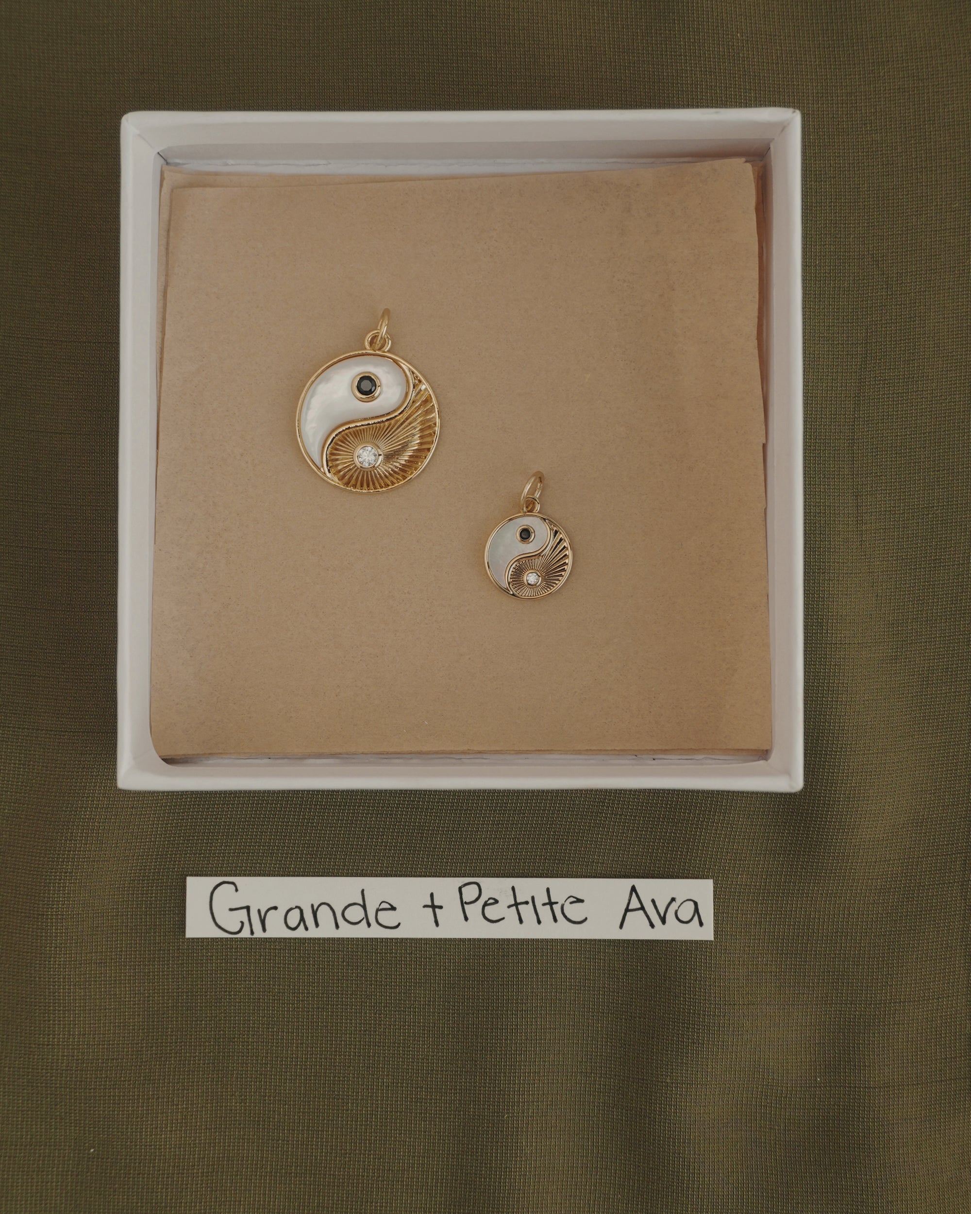 Two Mod + Jo Grande Ava Yin Yang charms, one large and one small 14k gold pendant, are displayed in a box on brown paper. Below the box is a label that reads "Grande + Petite Ava" on a green fabric background.