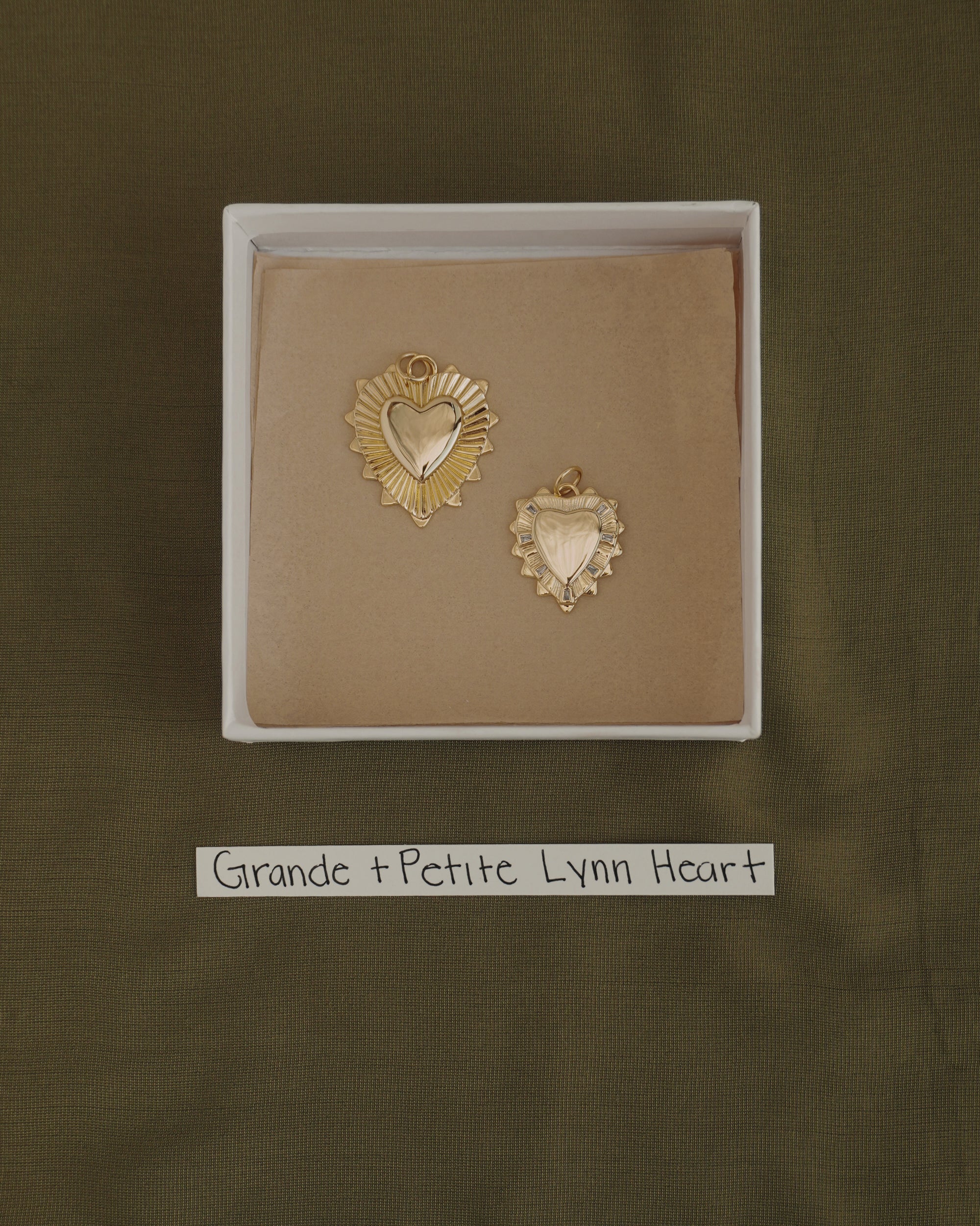 Two gold heart-shaped pendants, one larger and one smaller, are displayed in a box with a brown lining. Each pendant is crafted from 14k gold over brass by Mod + Jo, offering the perfect touch of everyday elegance. A label below reads "Grande + Petite Lynn Heart." The background features olive green fabric. The larger charm is the Grande Lynn Engravable Heart Charm.