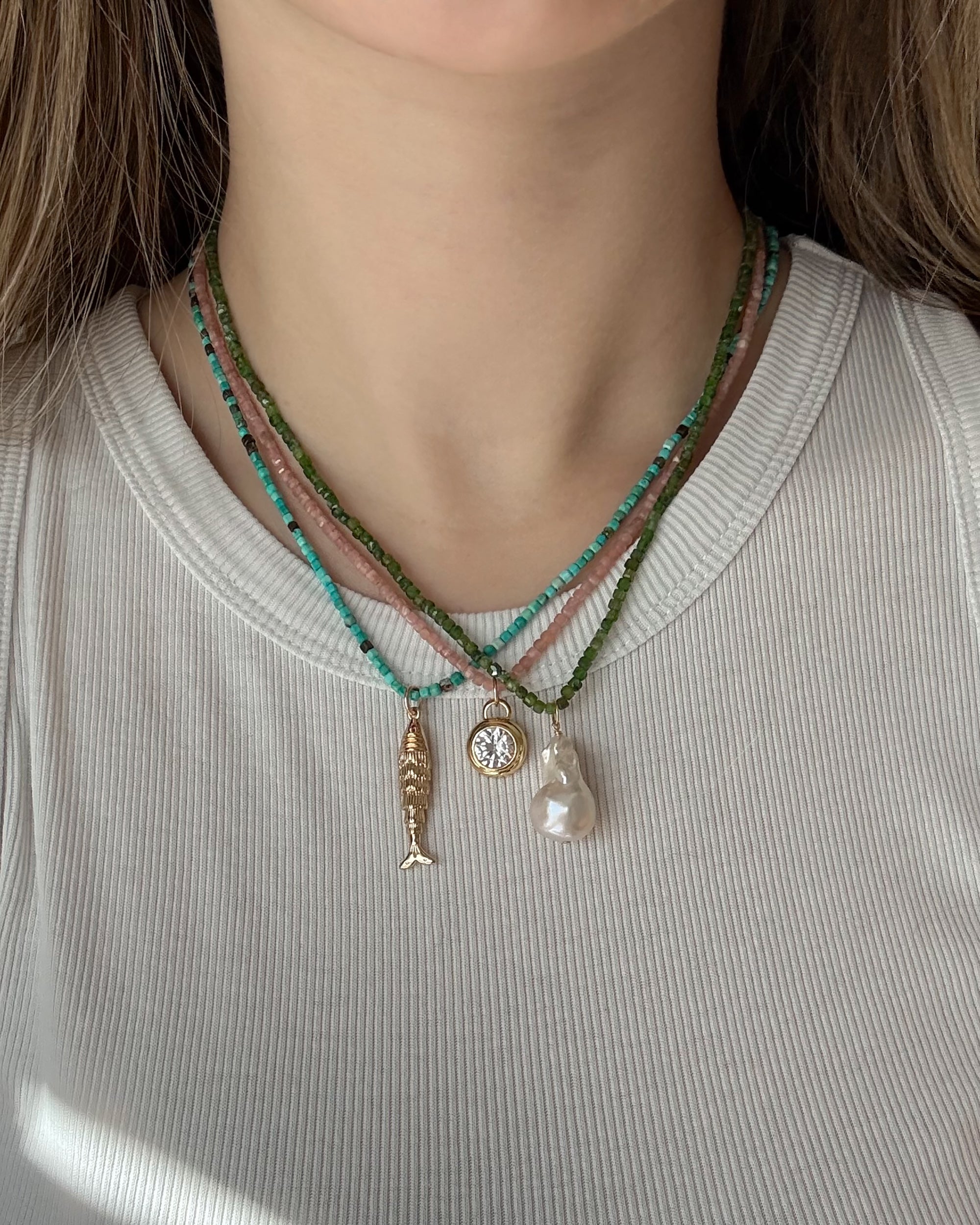 A person wears a white ribbed shirt and three layered Mod + Jo necklaces: one with green beads, one with turquoise beads, and one featuring the Jaci Baroque Pearl Charm—a 14k gold-plated fish charm, round charm, and large white baroque pearl.