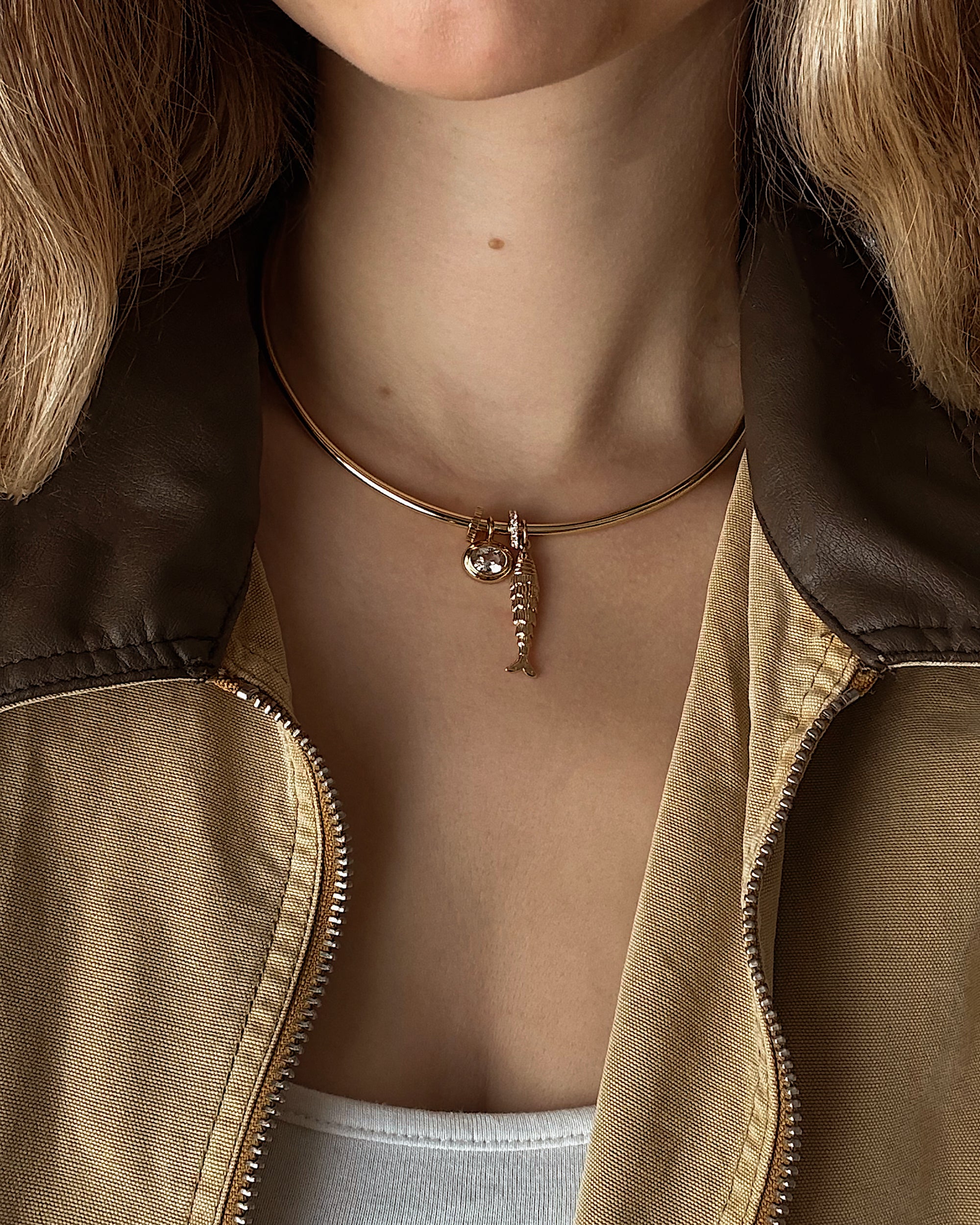 A person, shown from the shoulders down in a tan jacket with a brown collar and white shirt, wears a gold choker necklace featuring Mod + Jo’s Marla Sapphire Bezel Charm with lab-grown gemstones.