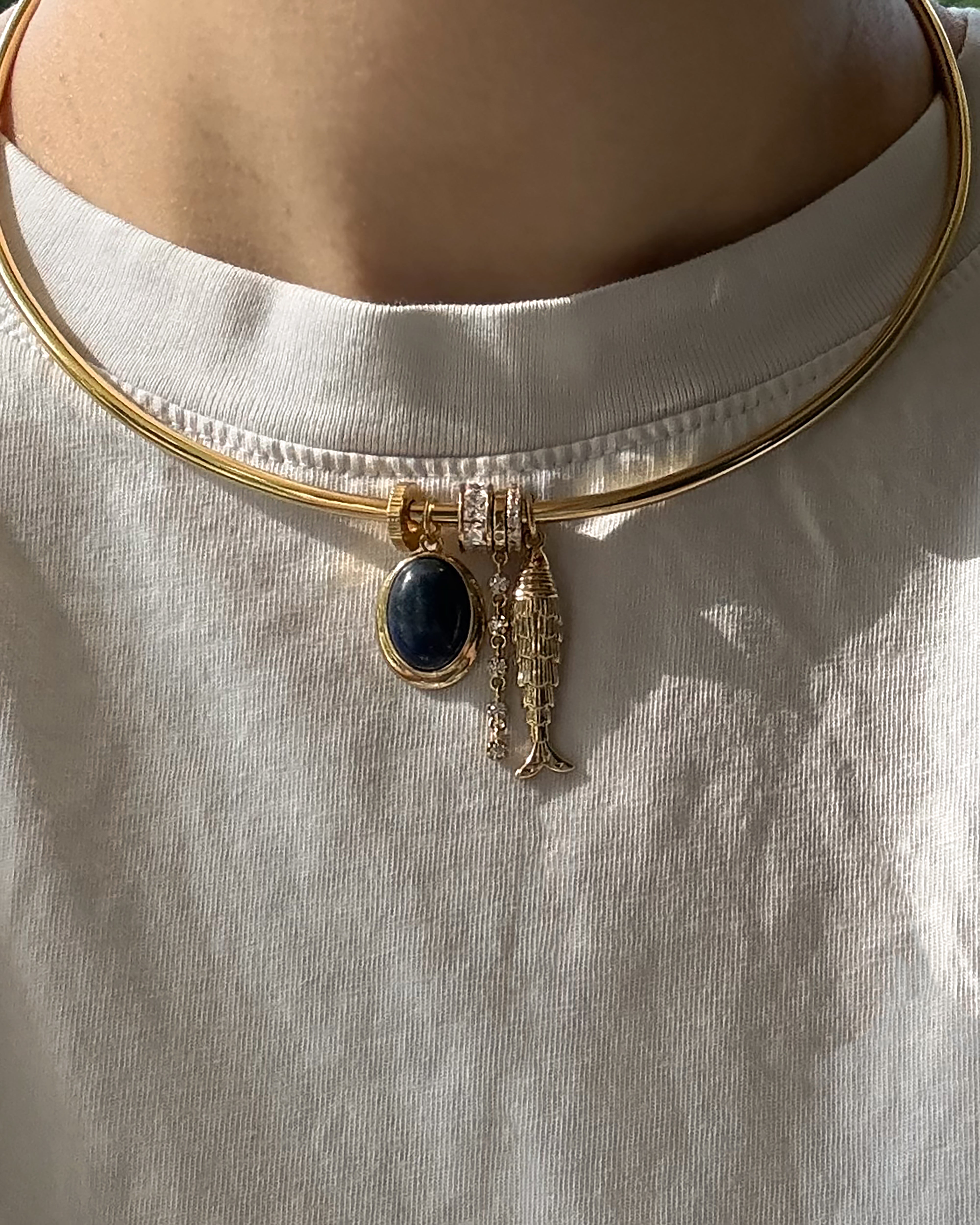 Worn over a white T-shirt, the Mod + Jo Demi Tennis Charm is a 14k gold plated choker necklace featuring a round dark blue stone charm and a gold fish-shaped pendant.