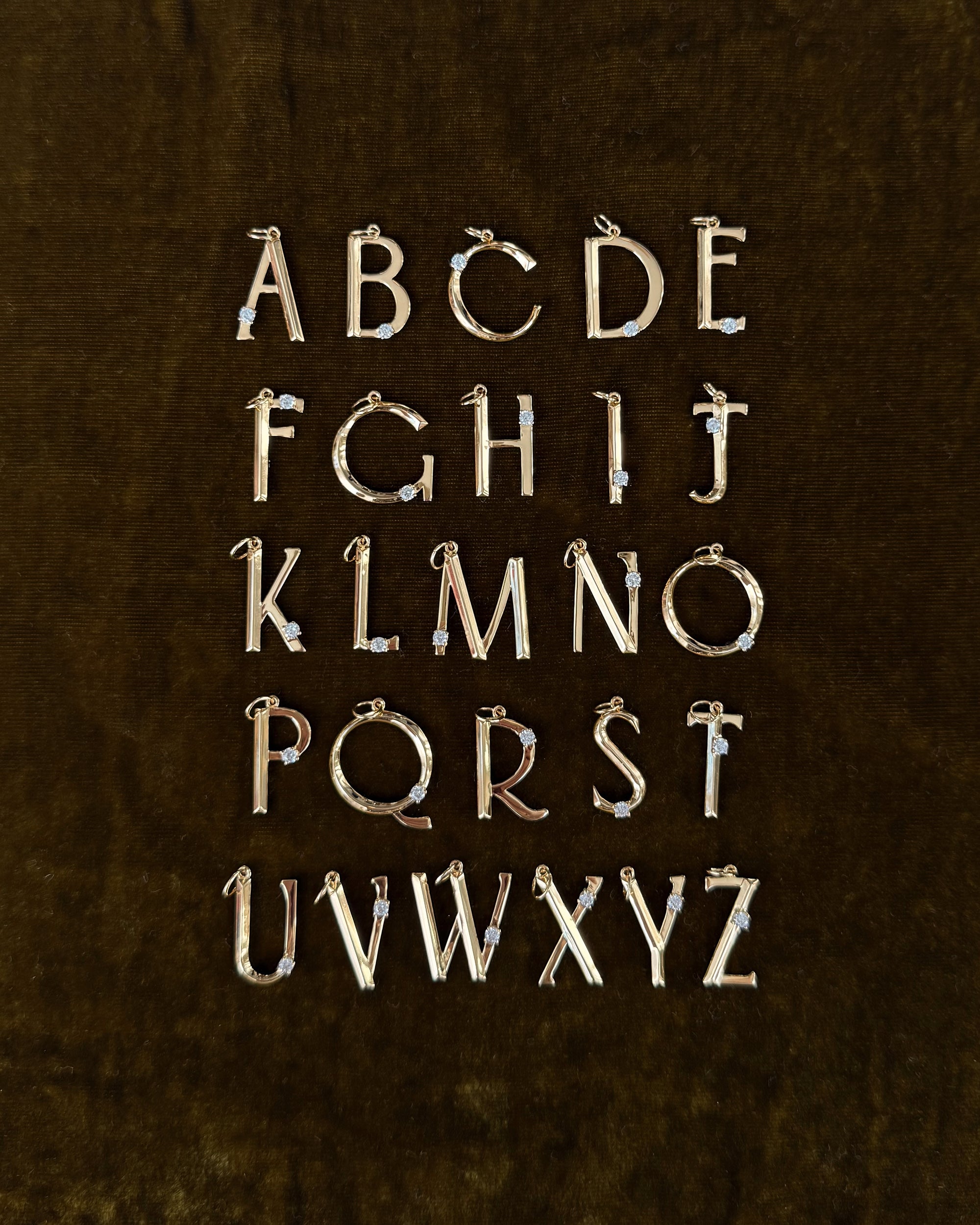 The Eloise Letter Charm by Mod + Jo features gold uppercase alphabet pendants with sparkling cubic zirconia accents, arranged in five rows on a dark brown velvet background, displaying A to Z.