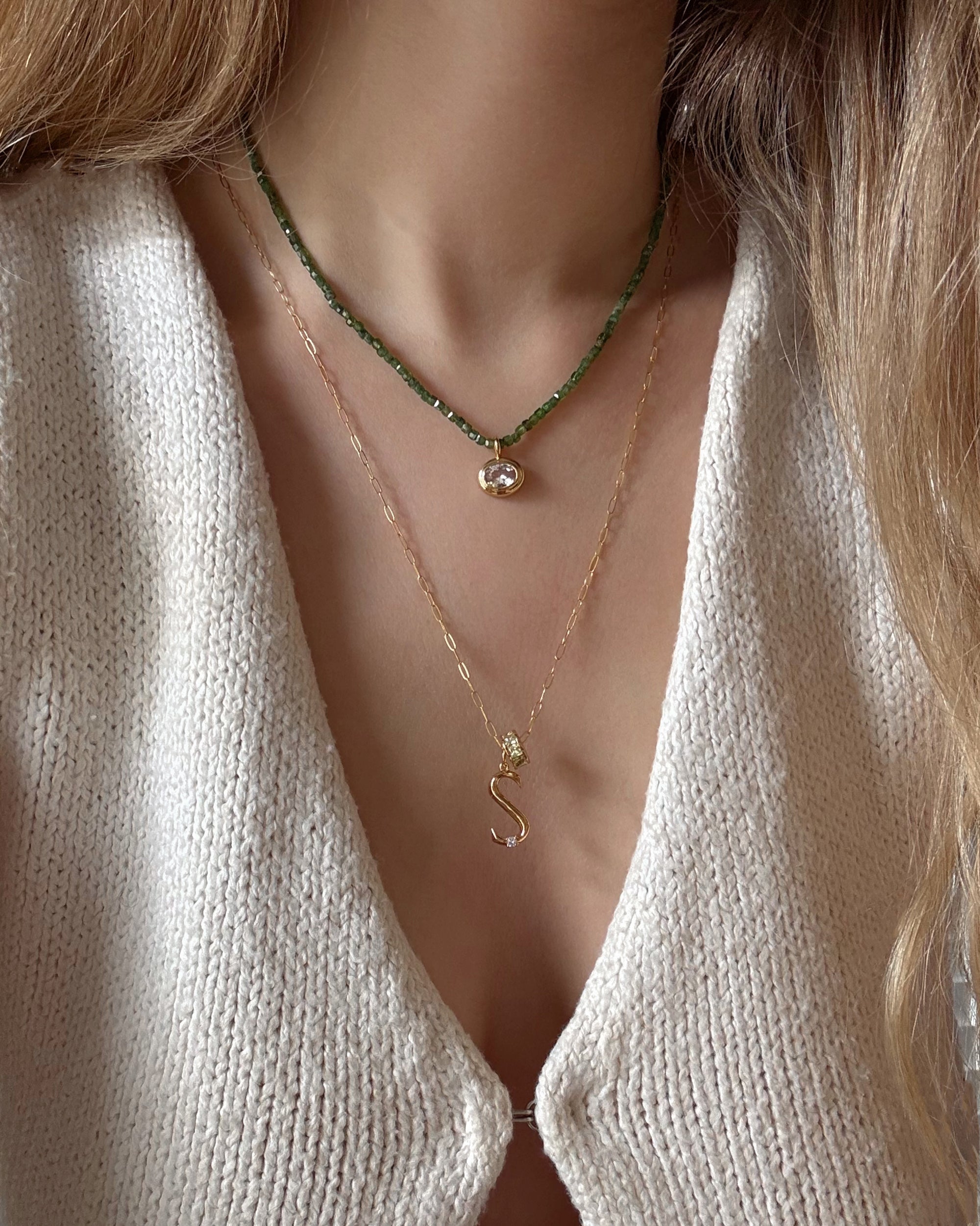 A woman in a cream knit top wears two layered Mod + Jo necklaces: a short green beaded necklace with an oval pendant and a longer 18k gold plated Eloise Letter Charm chain featuring an “S” gold charm.