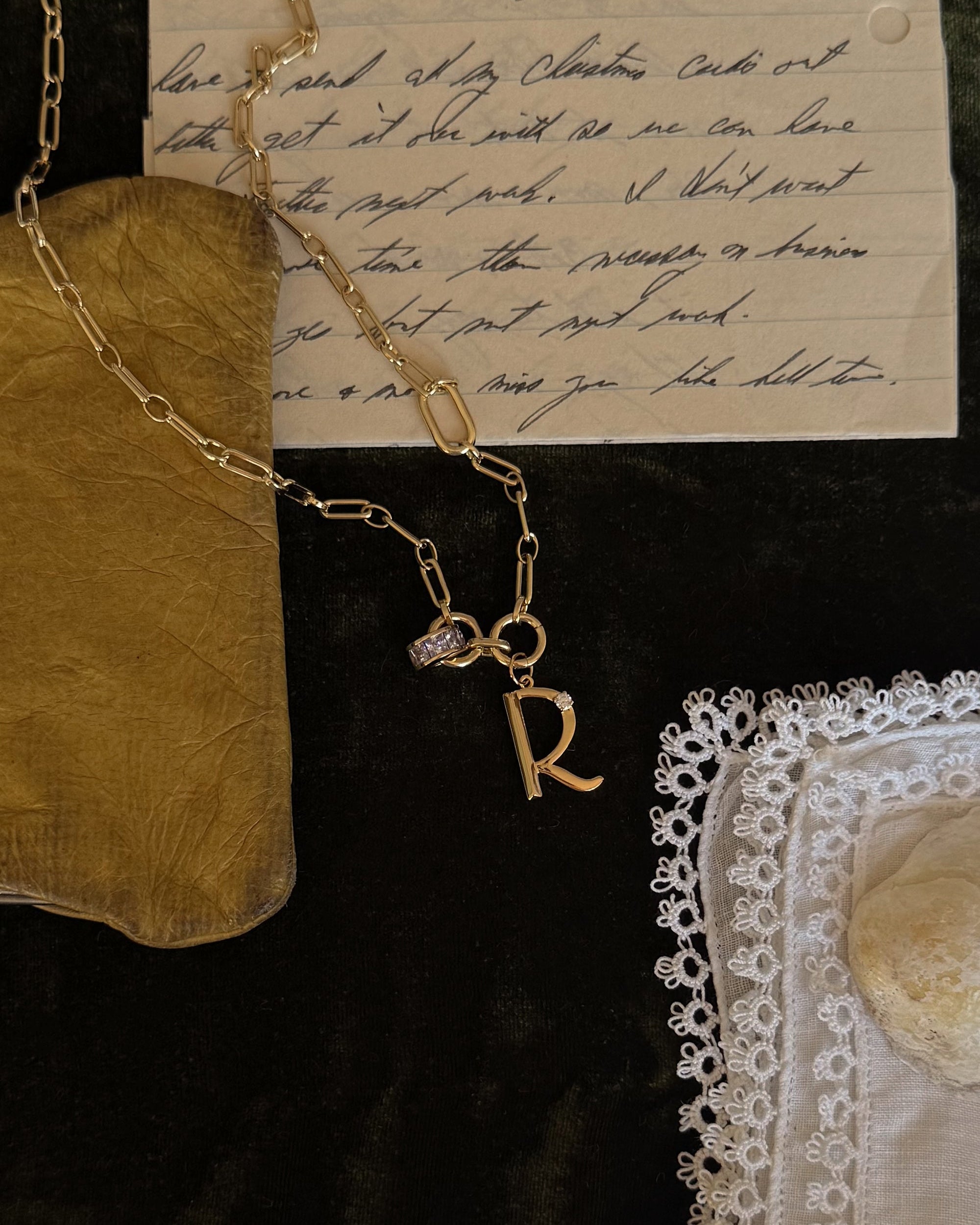 The Eloise Letter Charm necklace by Mod + Jo, featuring an "R" initial and small gem, is displayed on dark fabric beside a handwritten letter, golden pouch, and a white lace-trimmed cloth.