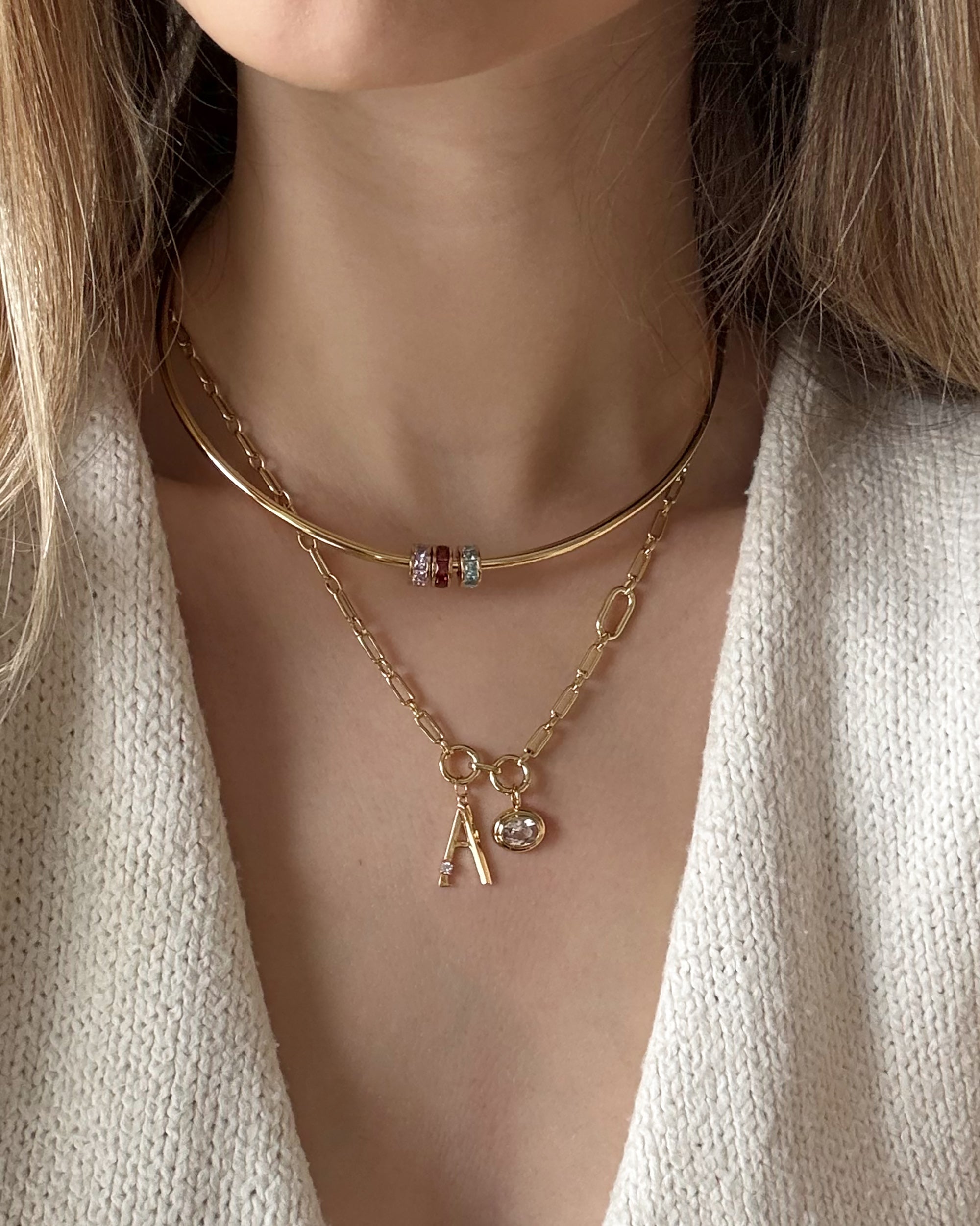 A woman wears two Mod + Jo necklaces: the Eloise Letter Charm with an "A" and a small cubic zirconia pendant, and another featuring colorful beads, styled over a cream textured top. The focus is on her neck and upper chest.