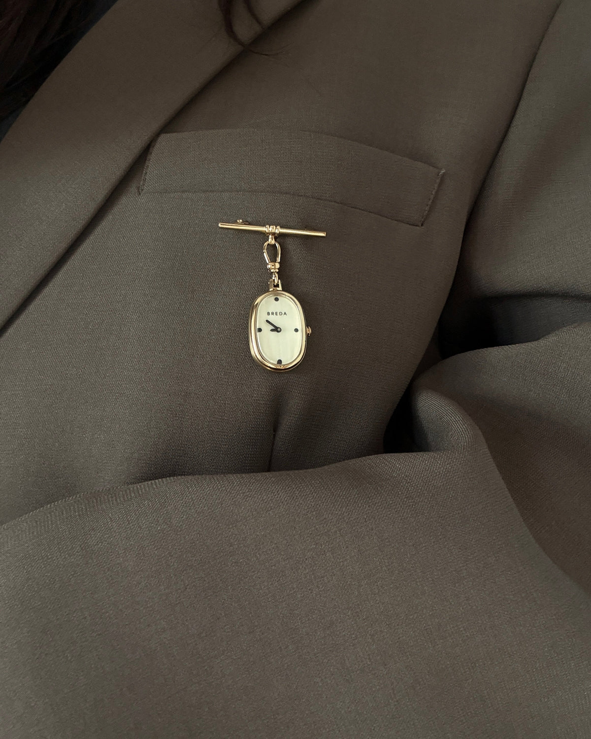 A gold-plated Jane Time Charm by BREDA featuring a mother of pearl dial, hanging from a horizontal pin attached to a dark blazer.