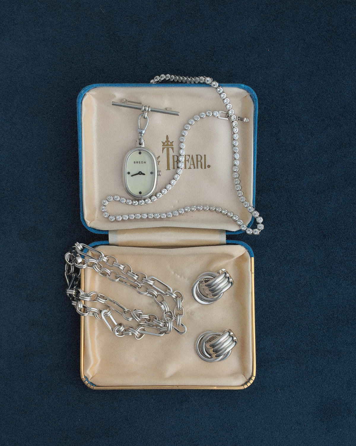 A Jane Time Charm by BREDA pendant watch with a silver chain, a rhinestone tennis bracelet, and two silver hoop earrings arranged inside a blue velvet-lined jewelry box.