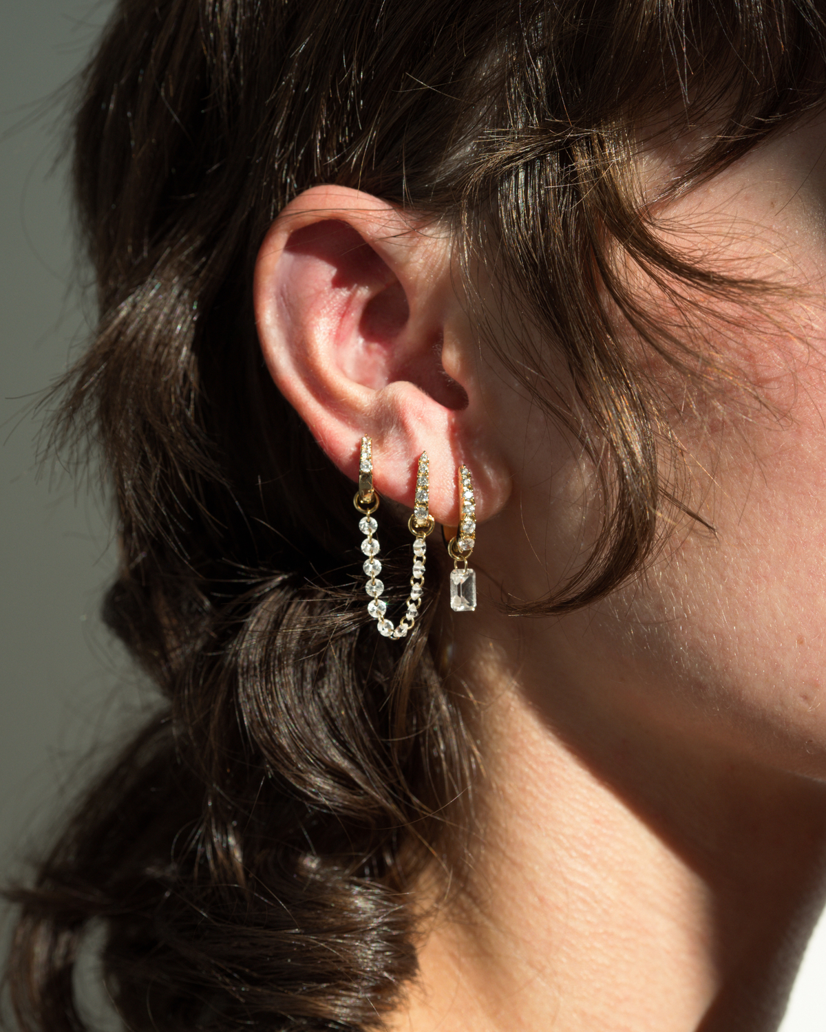 A person wearing Mod + Jo Iris Sapphire Earring Charms featuring two gold-plated hoops connected by a delicate chain of lab-grown white sapphires, with a single rectangular crystal charm hanging from the lower hoop.