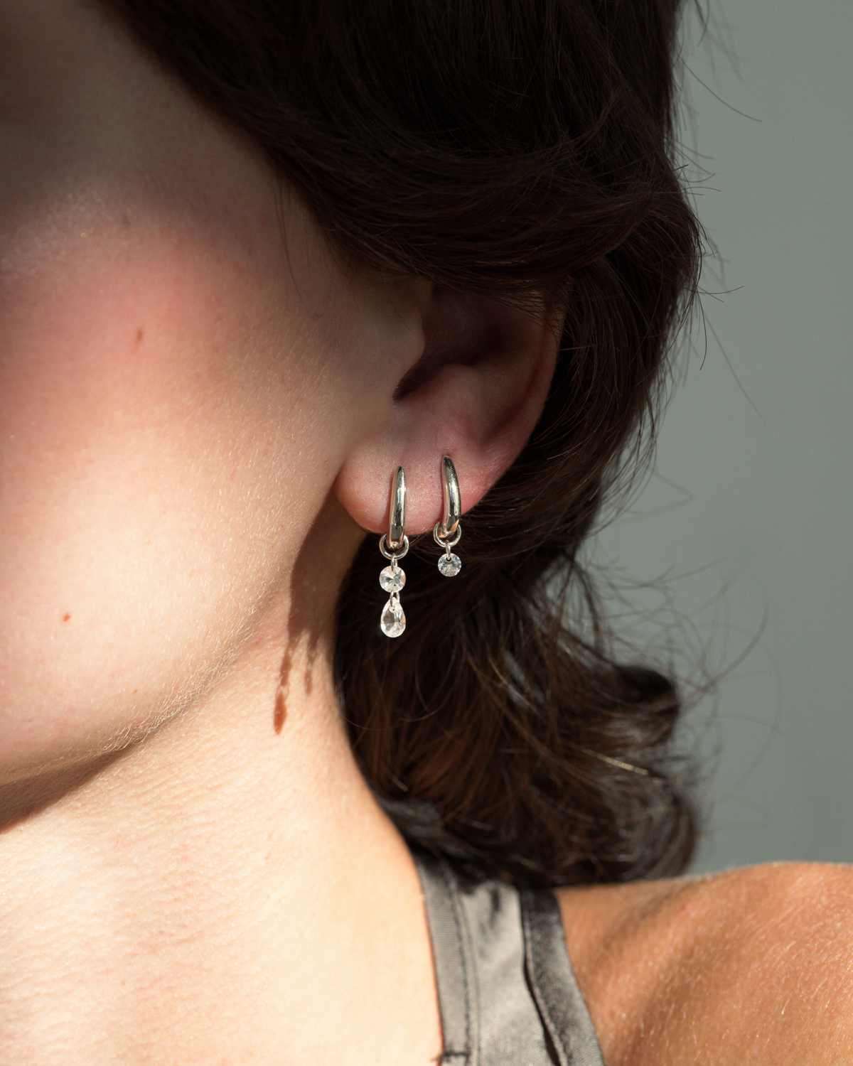 Mod + Jo Jules Smooth Huggies: two polished silver huggie earrings, each featuring a small dangling clear stone charm, worn in a pierced earlobe.