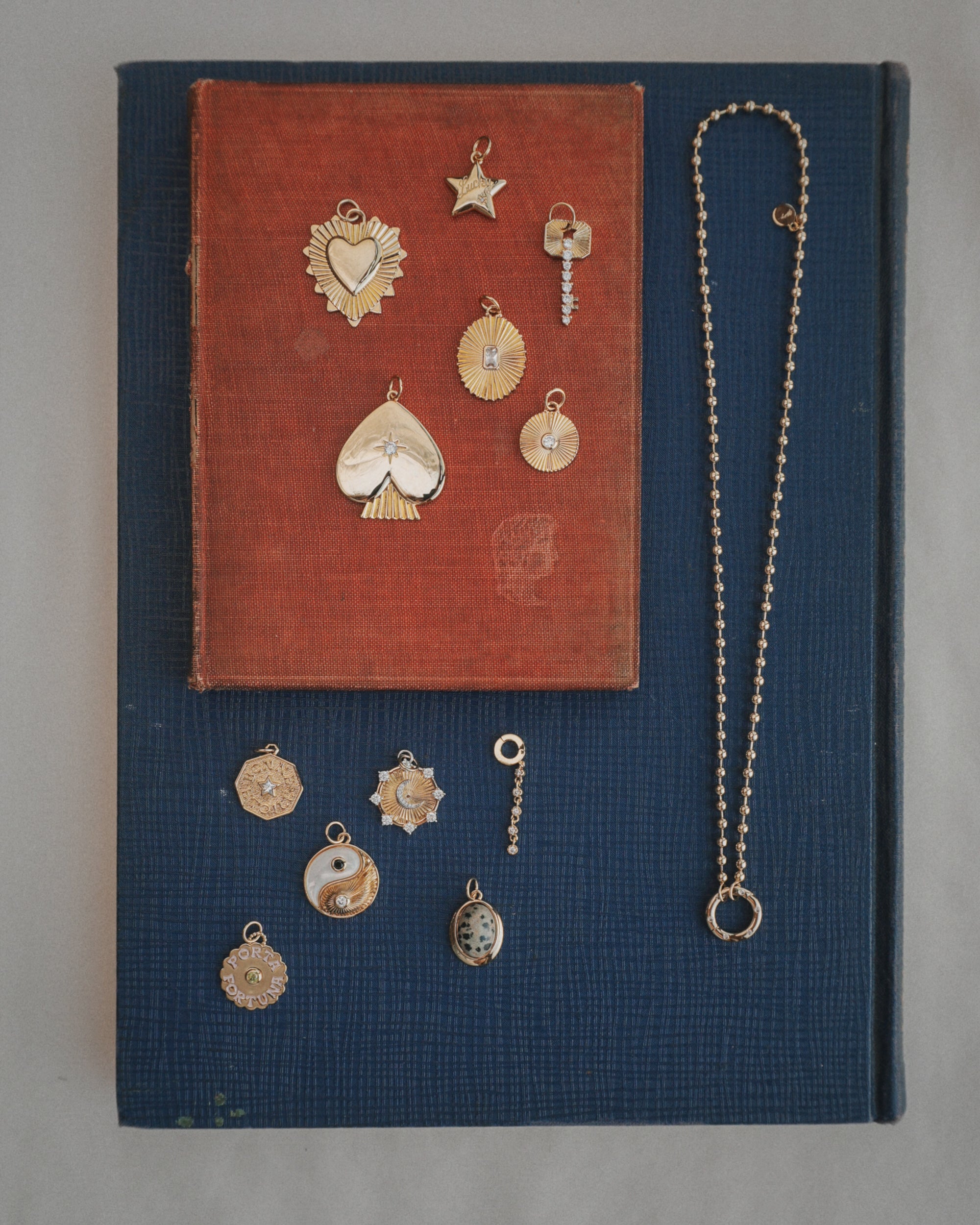 The Soren Ball Charm Clip Necklace by Mod + Jo is displayed on two stacked books, one red and one blue, adorned with gold charms in various shapes such as a heart, star, key, sun, yin-yang, and more—all arranged neatly for viewing.