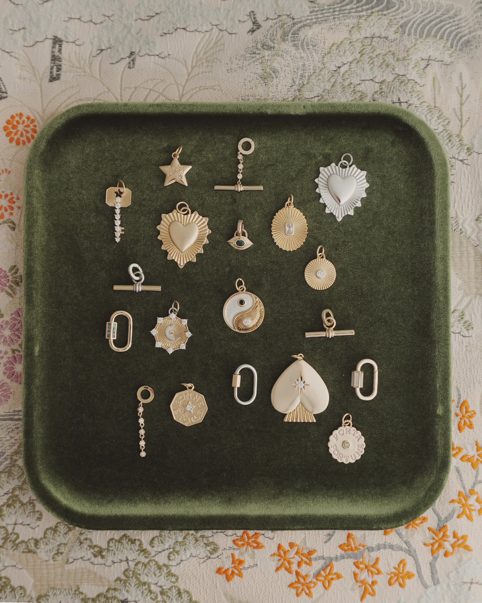 Nora Lock Carabiner charms by Mod + Jo—crafted in gold and silver finishes, featuring a range of shapes like hearts, stars, a yin-yang motif, and geometric forms (some embellished with natural semi-precious stones such as blue lace agate and highlighted with elegant 14k plating)—are artfully arranged on a dark green velvet tray. The backdrop is a richly patterned floral fabric.