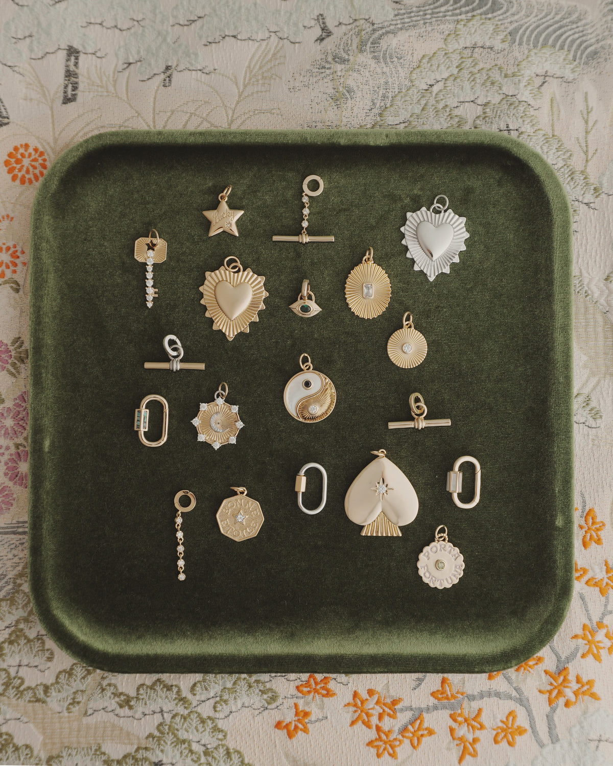 A green velvet tray presents 19 assorted gold and silver charms from Mod + Jo, including the elegant Grande Lynn Engravable Heart Charm, star motifs, yin-yang symbols, and geometric designs—all arranged neatly. The tray sits atop a floral-patterned fabric, accentuating the radiance of the collection’s 14k gold jewelry pieces.