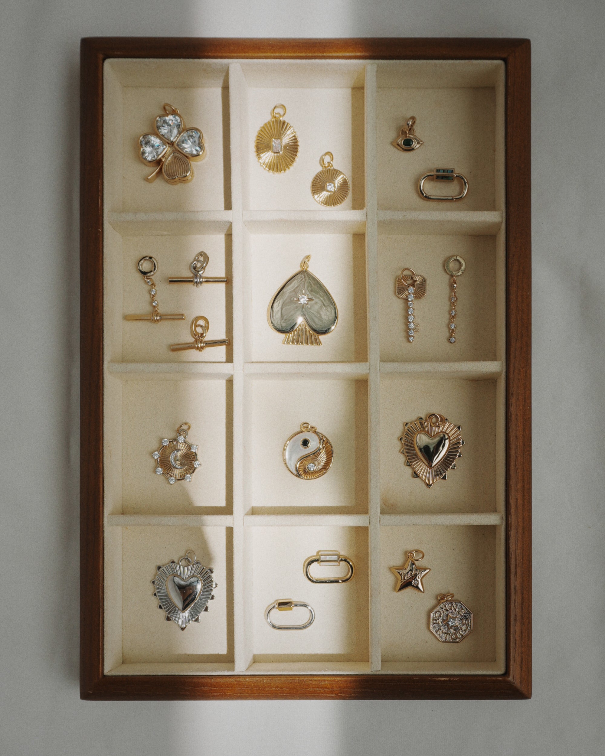 The Mod + Jo Cora Fluted Charm is displayed in a wooden jewelry box with twelve compartments, alongside gold and silver charms such as hearts, stars, an eye, a yin-yang symbol, and various geometric shapes—some highlighted with emerald-cut crystals—all arranged neatly on a cream fabric lining.