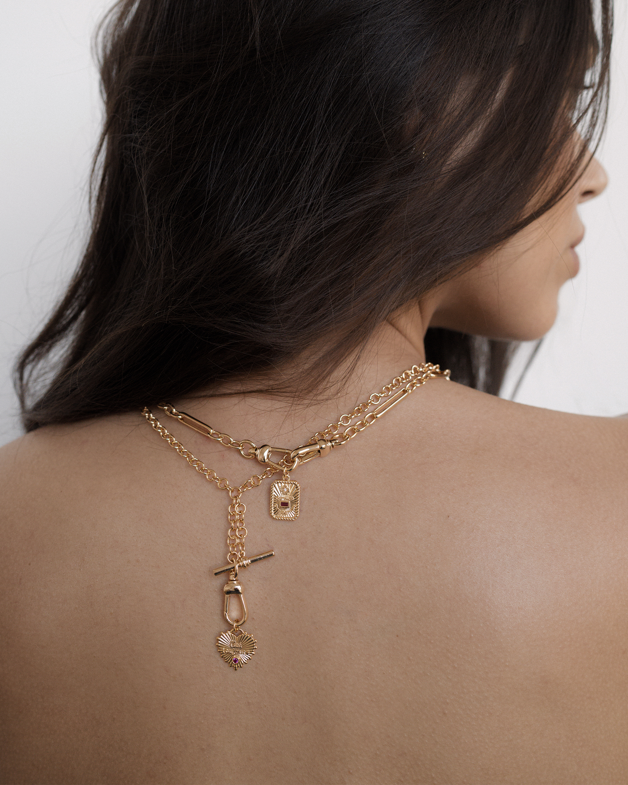 A woman with long, dark hair poses with her back to the camera, showcasing layered gold necklaces draped down her back with small pendants and charms. The background is neutral and minimal.