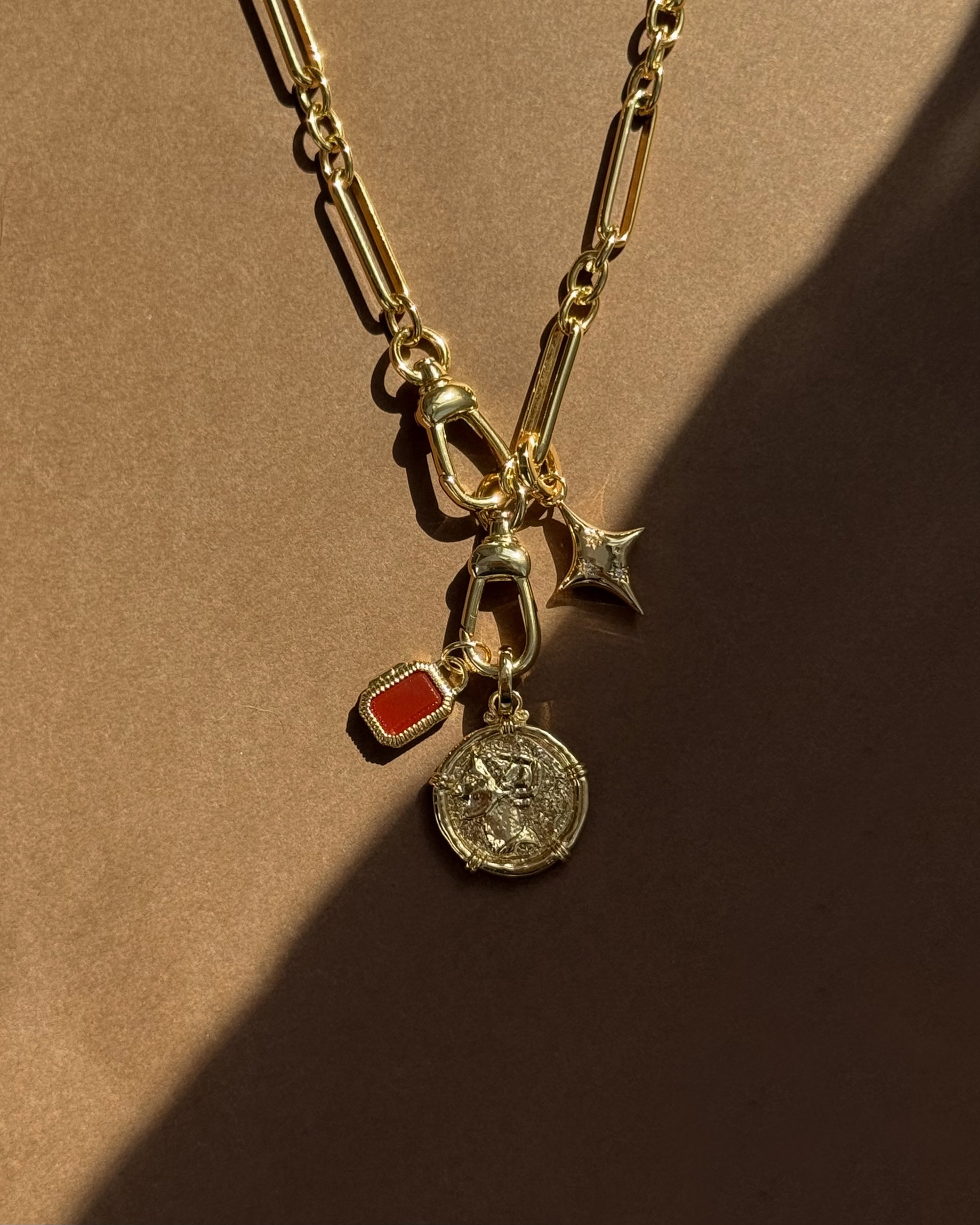 A Mod + Jo gold chain necklace with four charms—a red rectangle gem, a coin with a figure, a crescent moon, and the Celeste Puffy Star Charm—rests on a brown background with dramatic shadows.