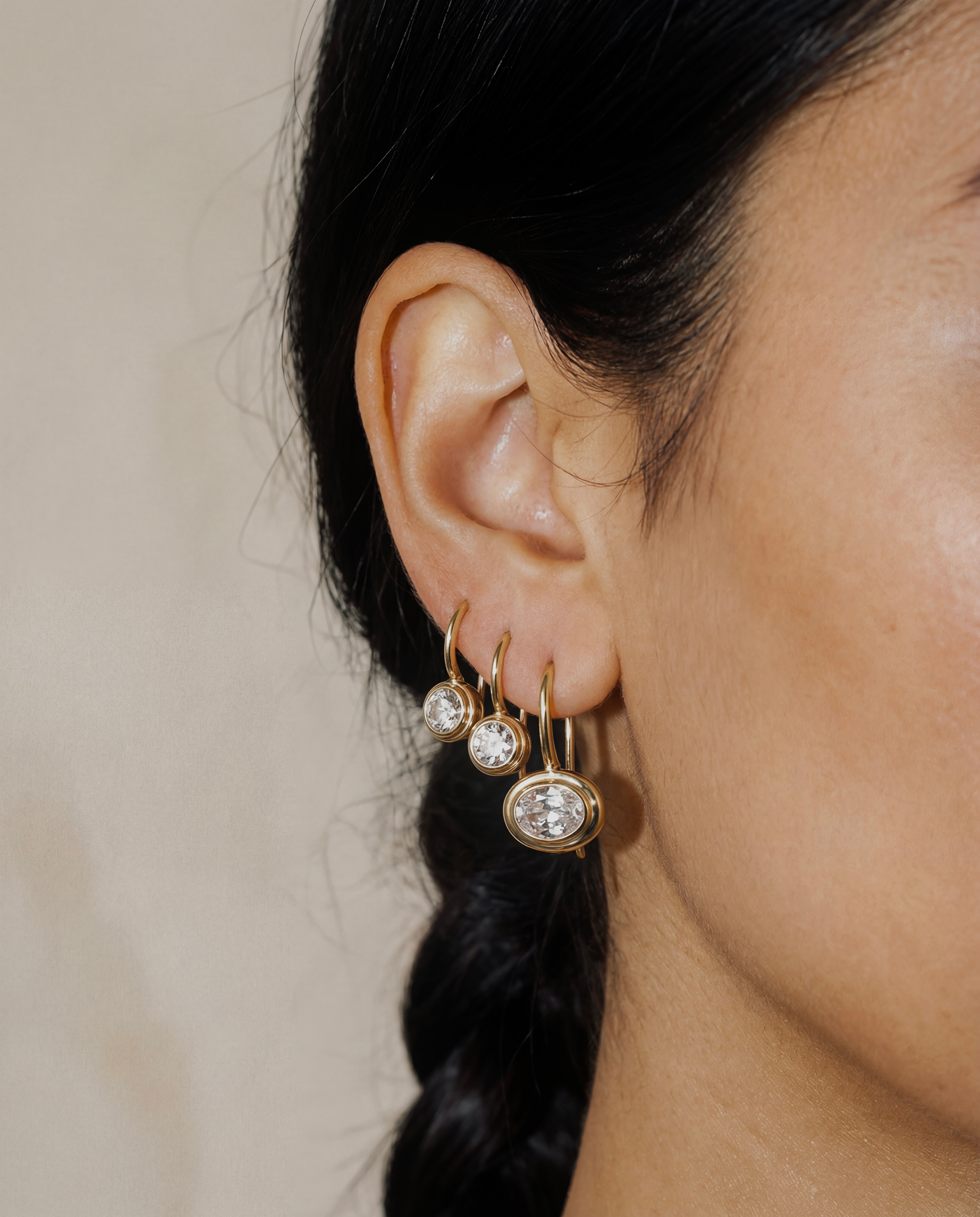 Close-up of an ear wearing gold hoop earrings with clear gemstones on a neutral background