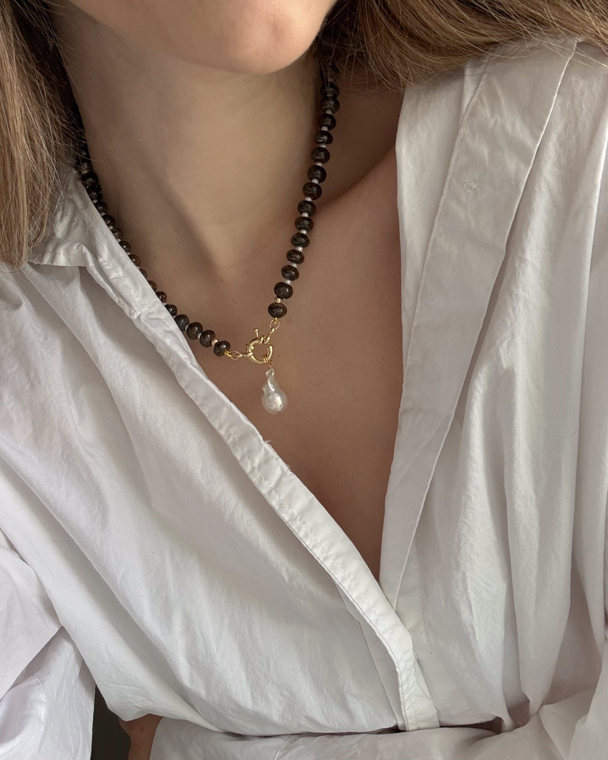 A person wears a white, open-collared blouse and the Mod + Jo Macey Gemstone Necklace, featuring genuine gemstones, a gold clasp, and a dangling pearl pendant for an elegant look.