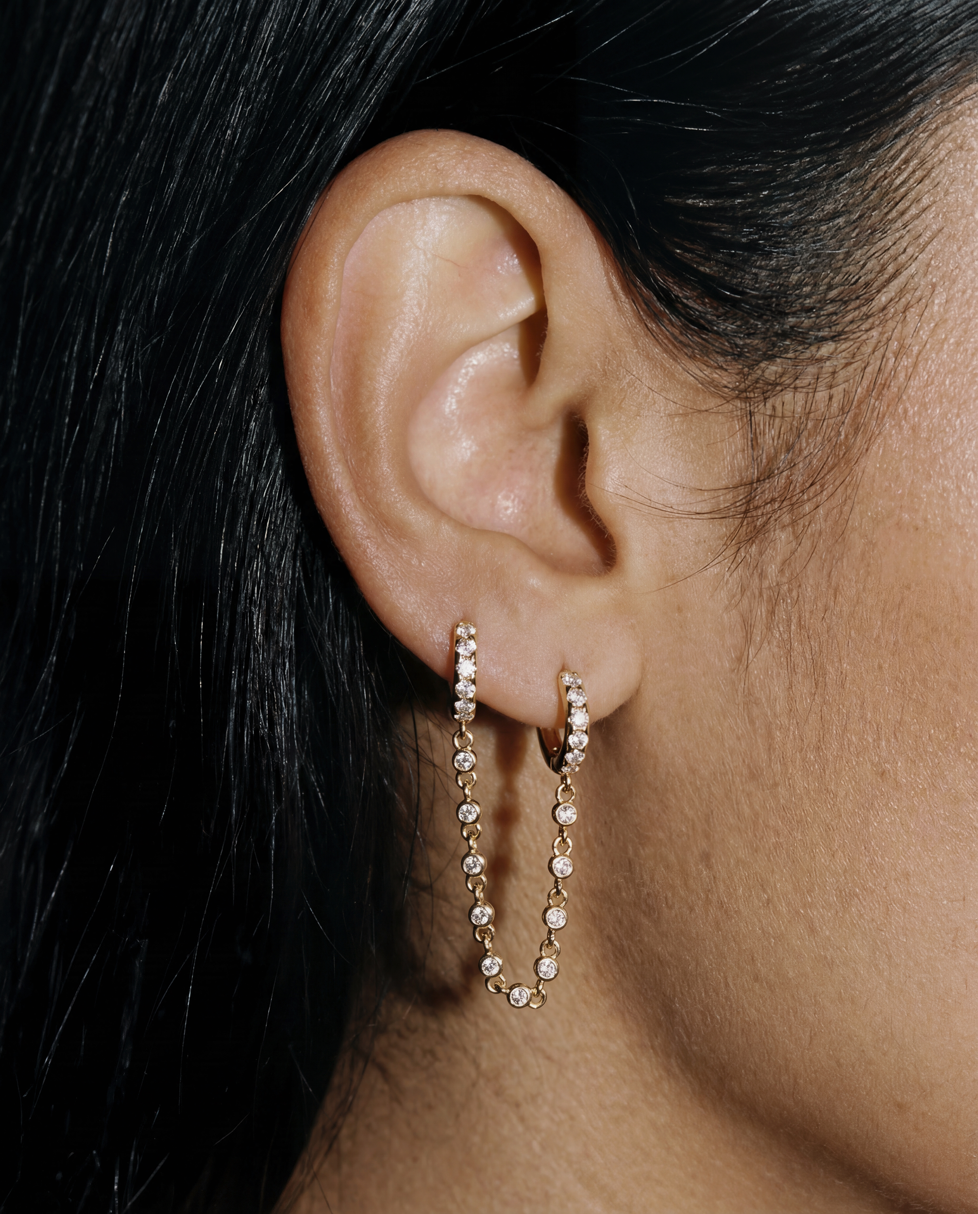 Close-up of an ear wearing gold hoop earrings with small czs and a  bezel chain connecting the two.