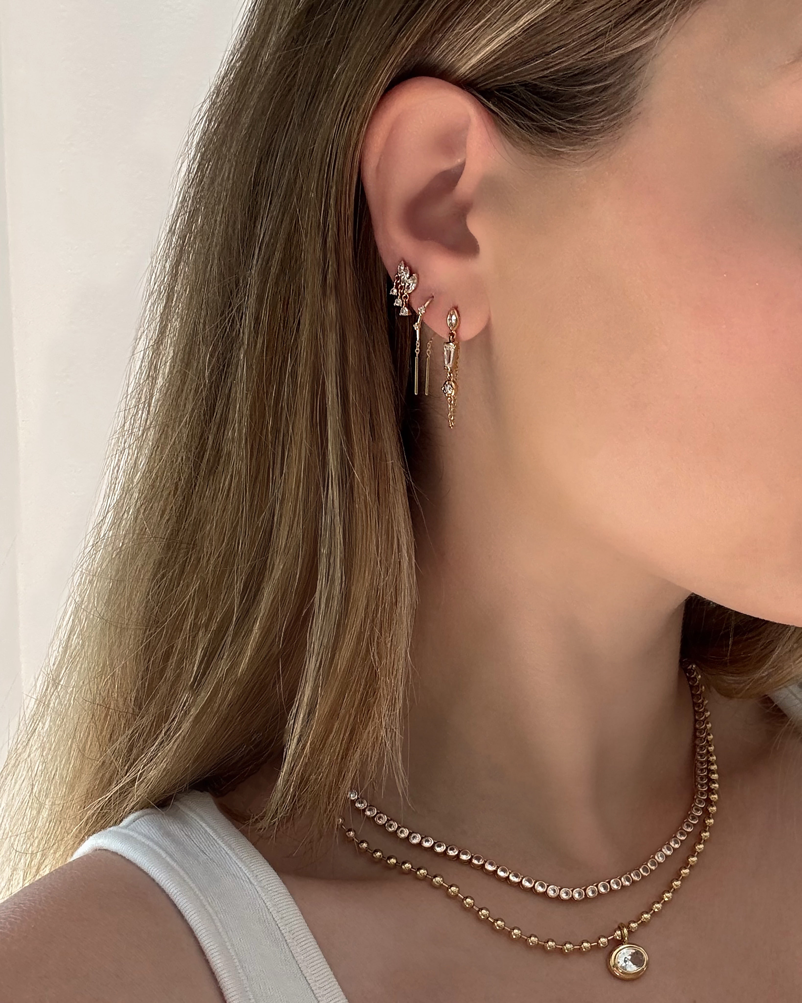 Close-up of a woman wearing the Mod + Jo Gabby Marquise Dangle Studs along with two gold necklaces. She has straight blonde hair and is dressed in a white tank top, set against a softly lit, plain background.