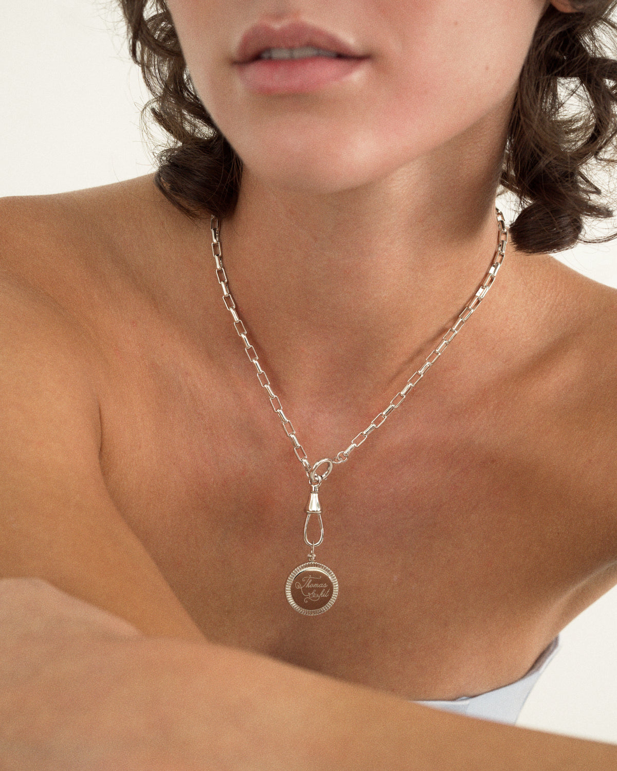 A person wearing the Gia Engravable Charm, featuring a silver chain necklace with an engravable, coin-edge disc pendant hanging from a decorative clasp.