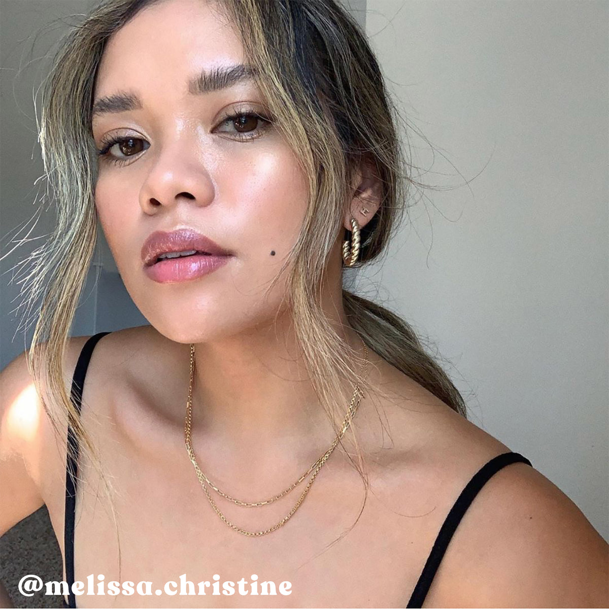 A woman with glowing skin, soft wavy hair, gold hoop earrings, and layered gold necklaces poses in a black strap top. Her makeup is natural and dewy. The Instagram handle @melissa.christine appears in the lower left corner.