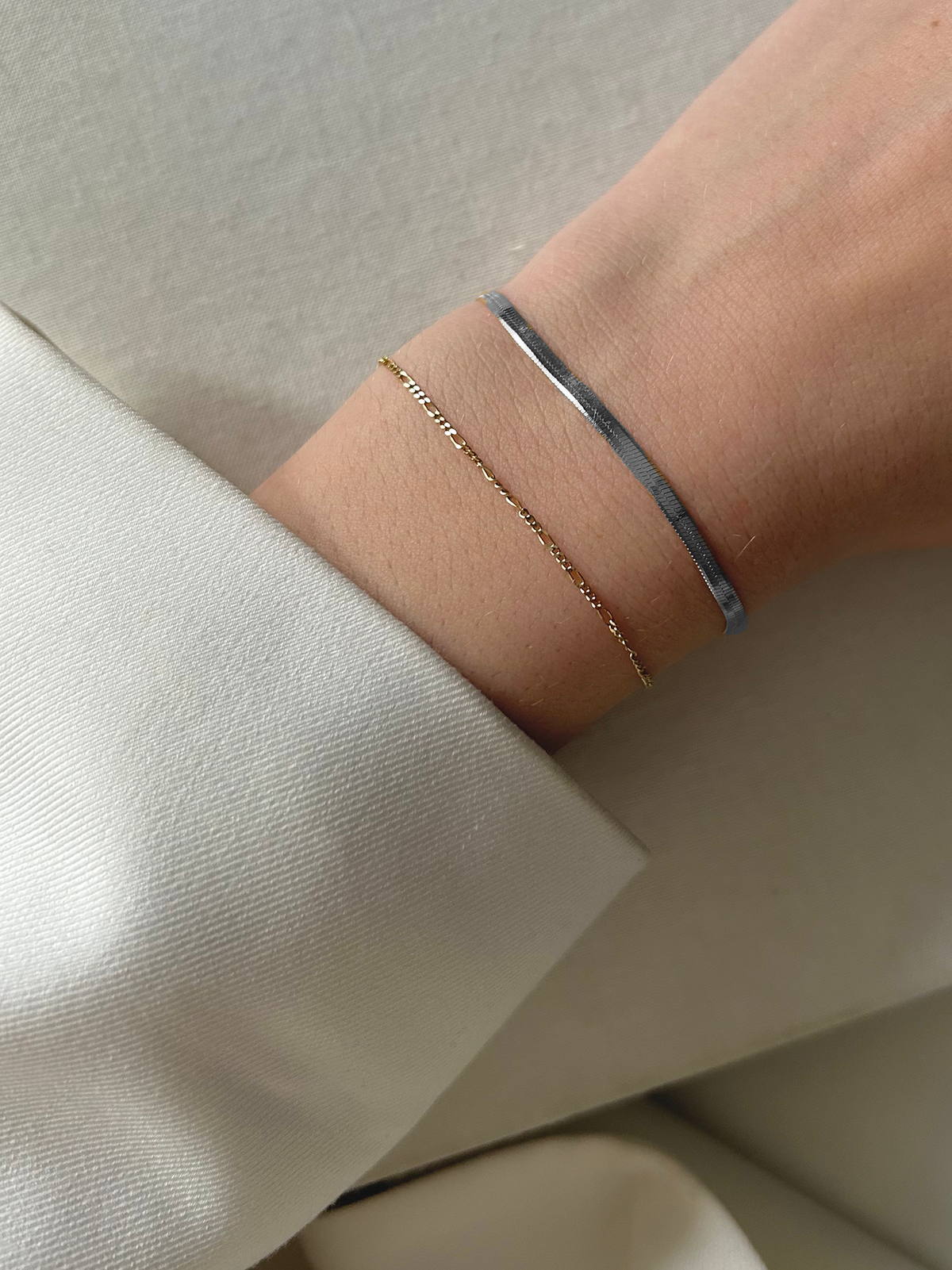 A close-up of a wrist wearing a thin gold chain bracelet and a sleek silver bangle, with a white sleeve partially visible. The background is a soft, textured light fabric.