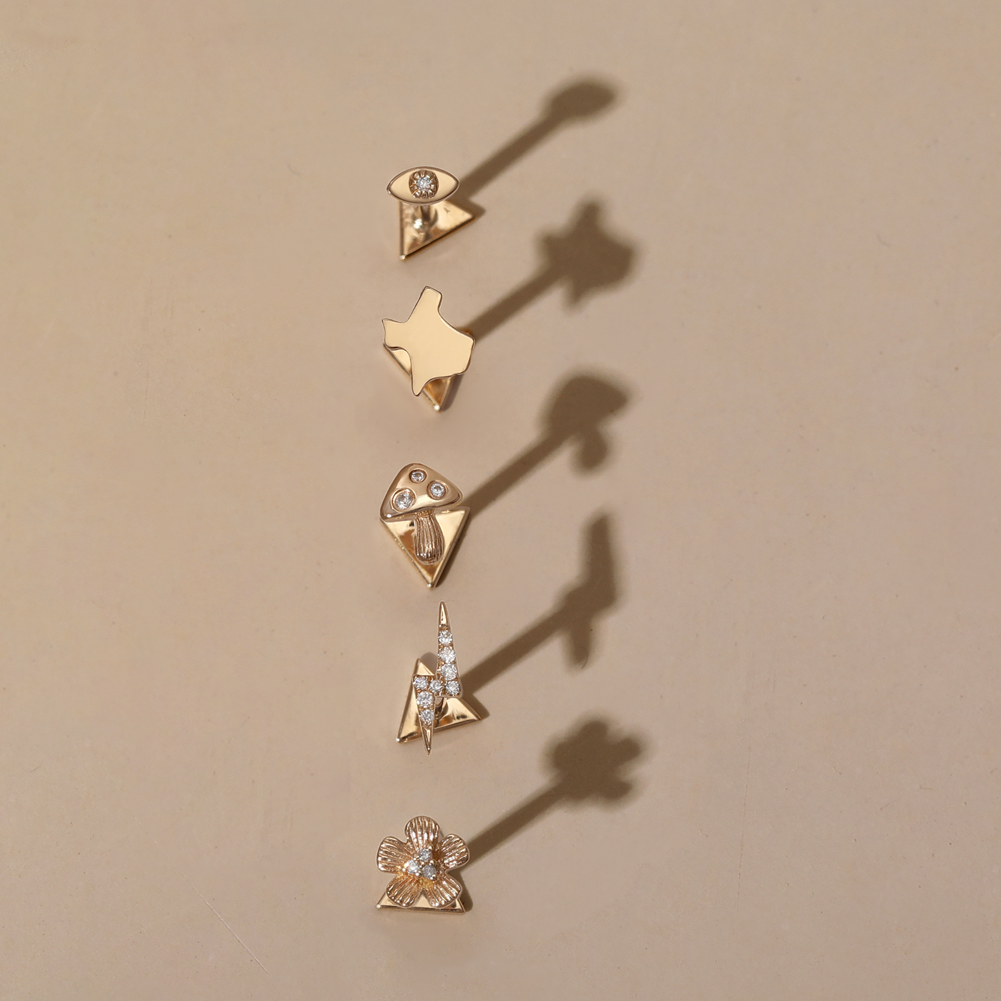 Five unique gold pins with different shapes and small gemstones are arranged vertically on a tan surface, each casting distinct shadows to the right.