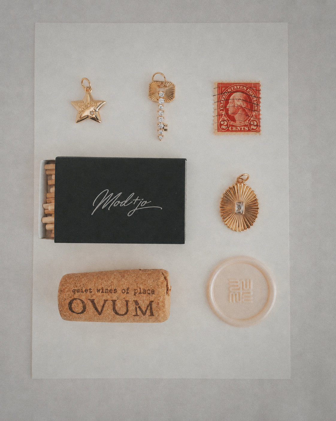 A flat lay featuring the Mod + Jo Noah Key Charm, alongside a red stamp, a black matchbox, a wine cork labeled OVUM, and a round beige item, all neatly arranged on a white background.