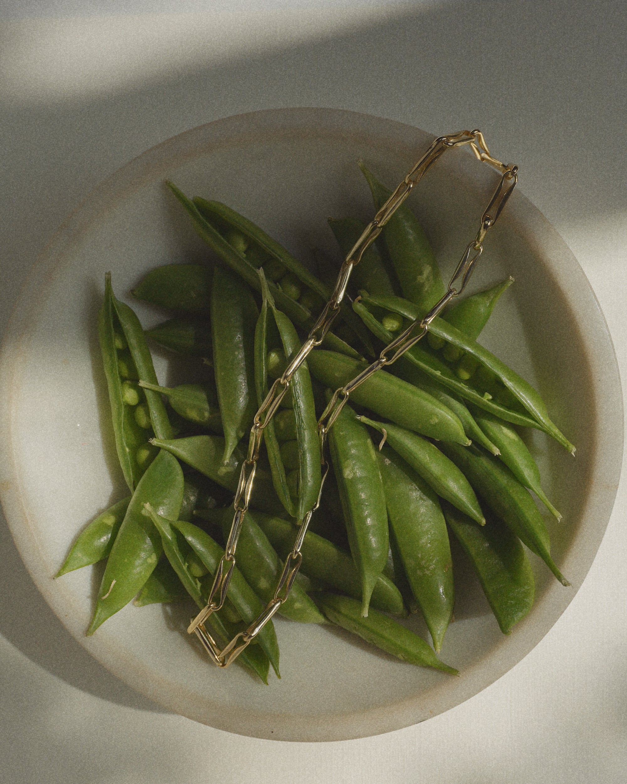 The Molly Elongated Link Chain Necklace by Mod + Jo, featuring a gold plate and elongated links, rests gracefully atop fresh green pea pods in a white bowl on a light-colored surface, illuminated by soft natural light and gentle shadows.